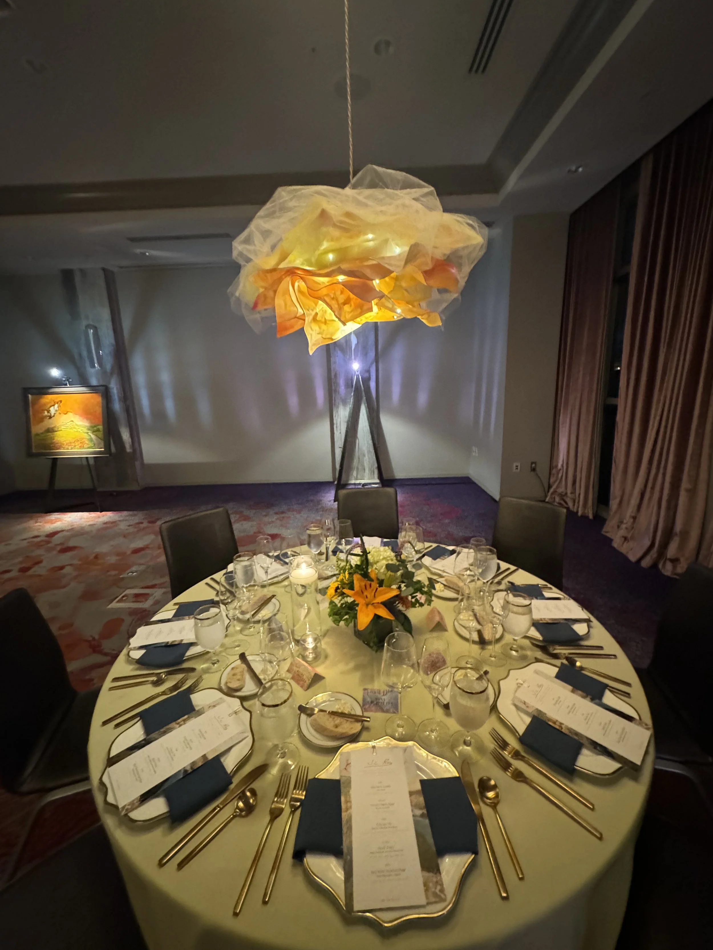 Round dinner table set with gold utensils, blue napkins, white plates, glasses, and floral centerpiece, in an elegant banquet hall with a hanging art piece and framed painting.