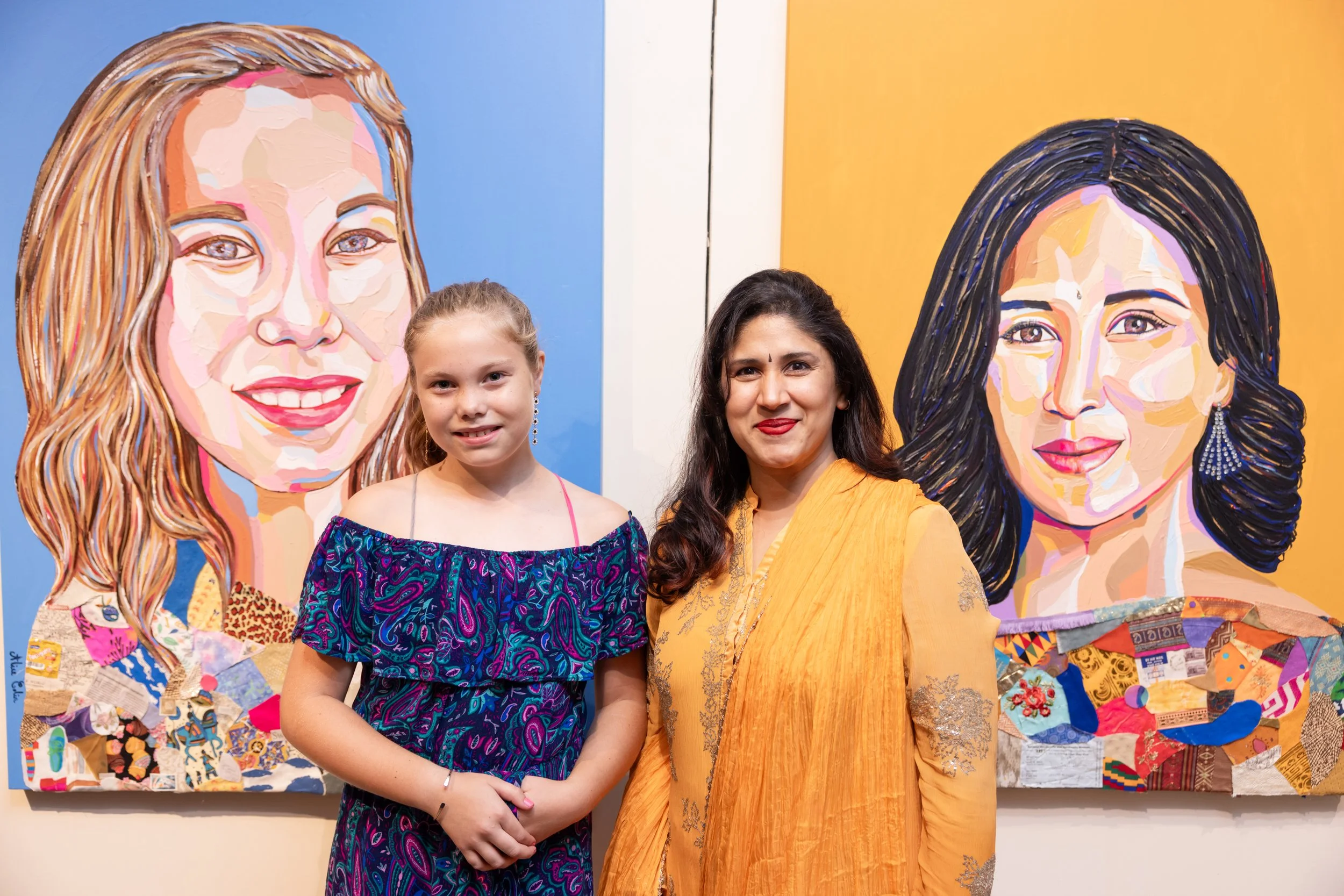 Two women, one young girl and one older woman, standing in front of colorful portraits of women painted in a collage style, with the woman on the right wearing a yellow-orange traditional outfit and the girl wearing a colorful patterned dress.