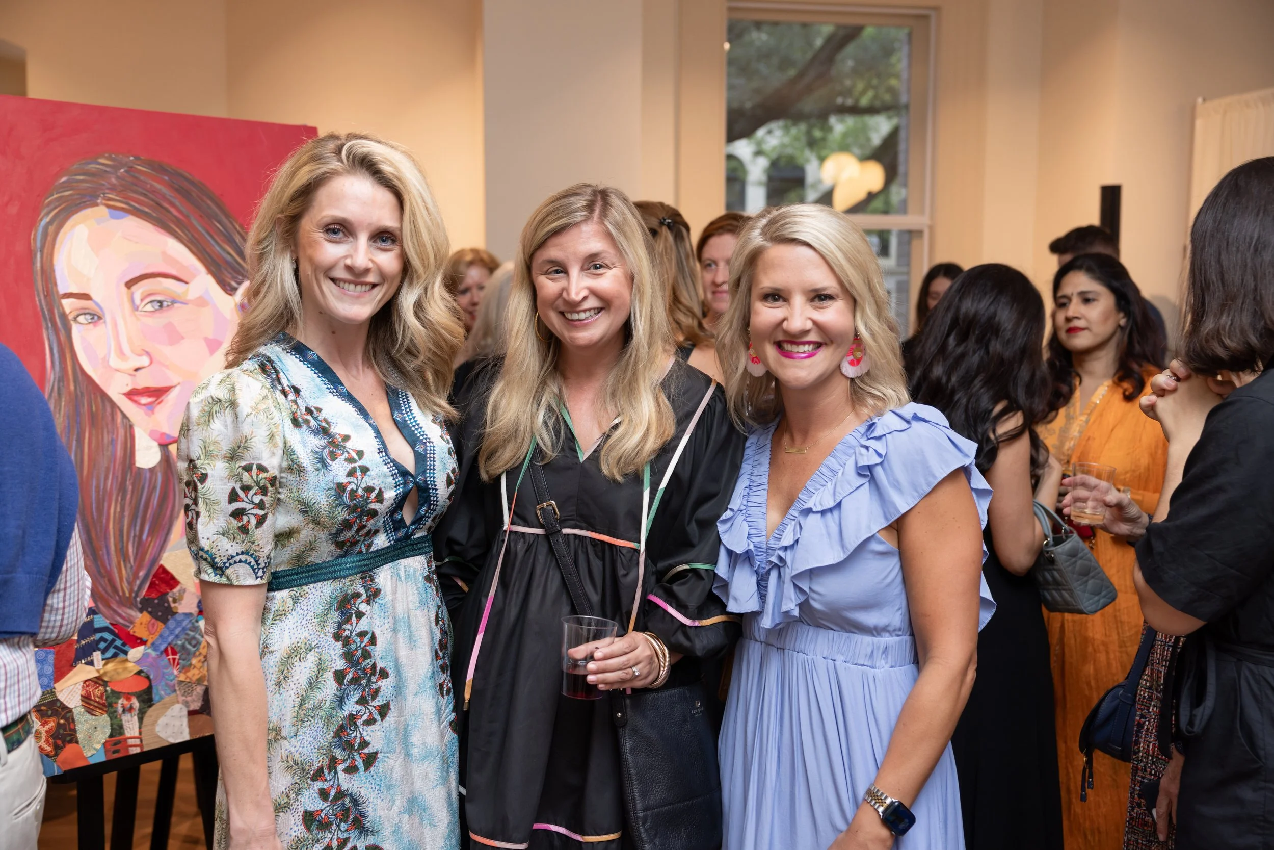 Group of women smiling at an art gallery event, with colorful artwork in the background and a window showing trees outside.