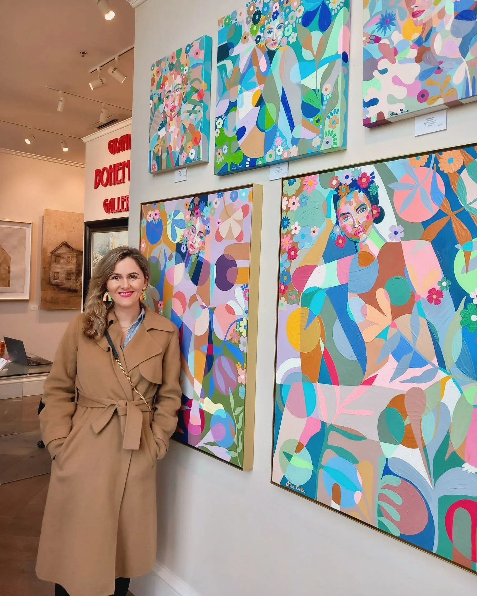 A woman with long, light brown hair wearing a beige trench coat, standing in an art gallery with colorful abstract portraits of women on the wall behind her.