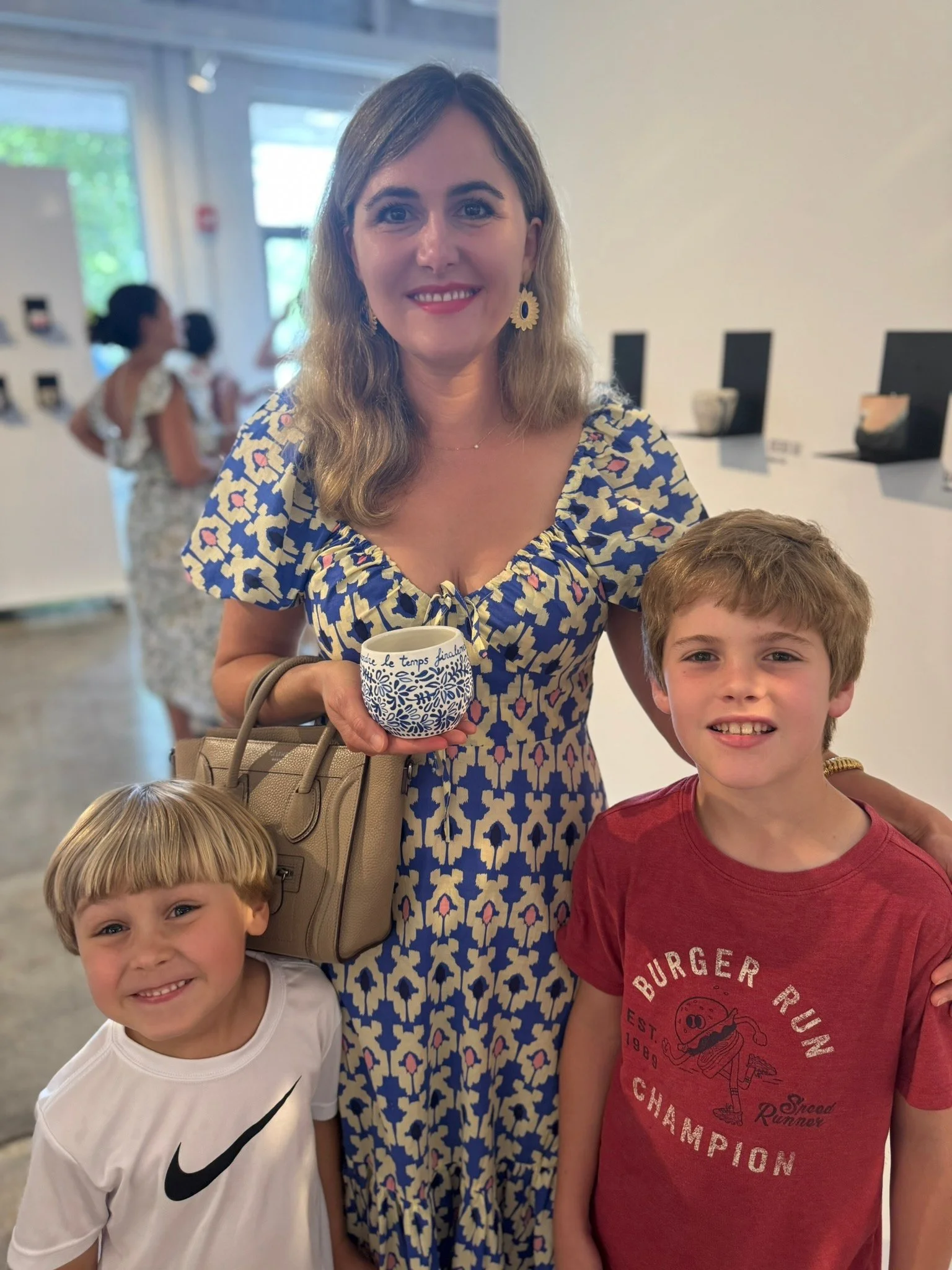 A woman with shoulder-length wavy hair, wearing a blue and yellow dress with a floral pattern and gold hoop earrings, holding a patterned white mug, standing with three young boys in a bright indoor setting.