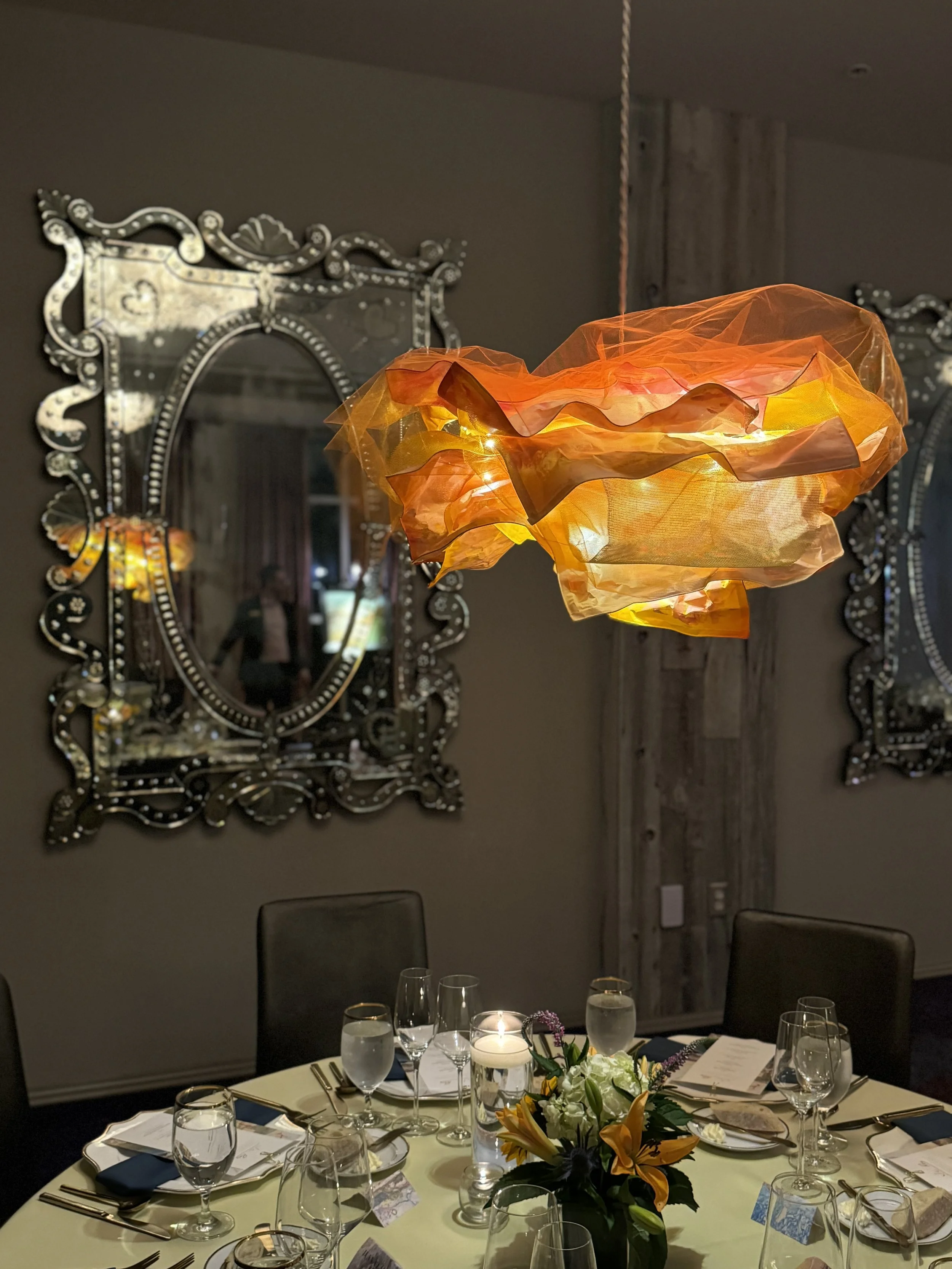 A round dining table set for a formal dinner with multiple glasses, utensils, napkins, and a floral centerpiece. Above is a modern, orange fabric pendant light. Two ornate silver mirrors with intricate frames hang on the wall behind.