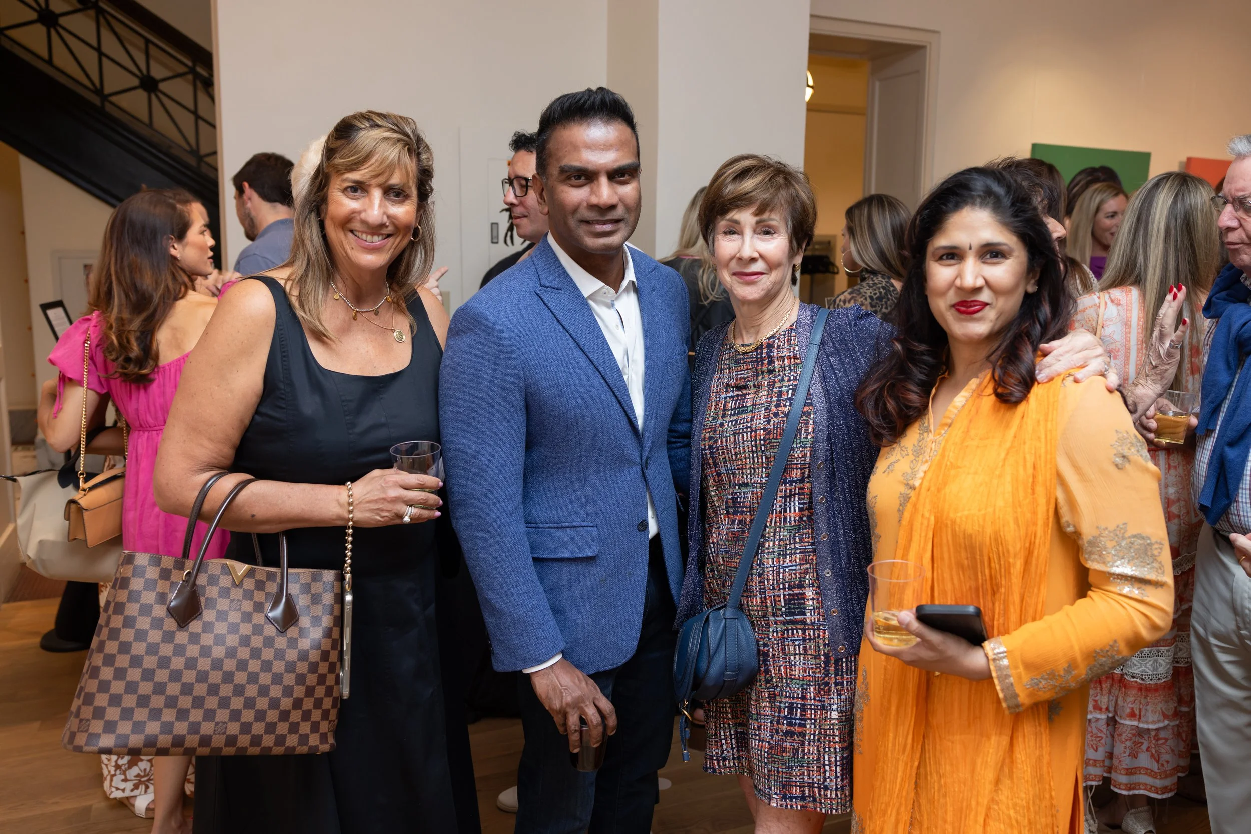 People at a social gathering, smiling and holding drinks, with a diverse group of individuals in a warmly decorated indoor setting.