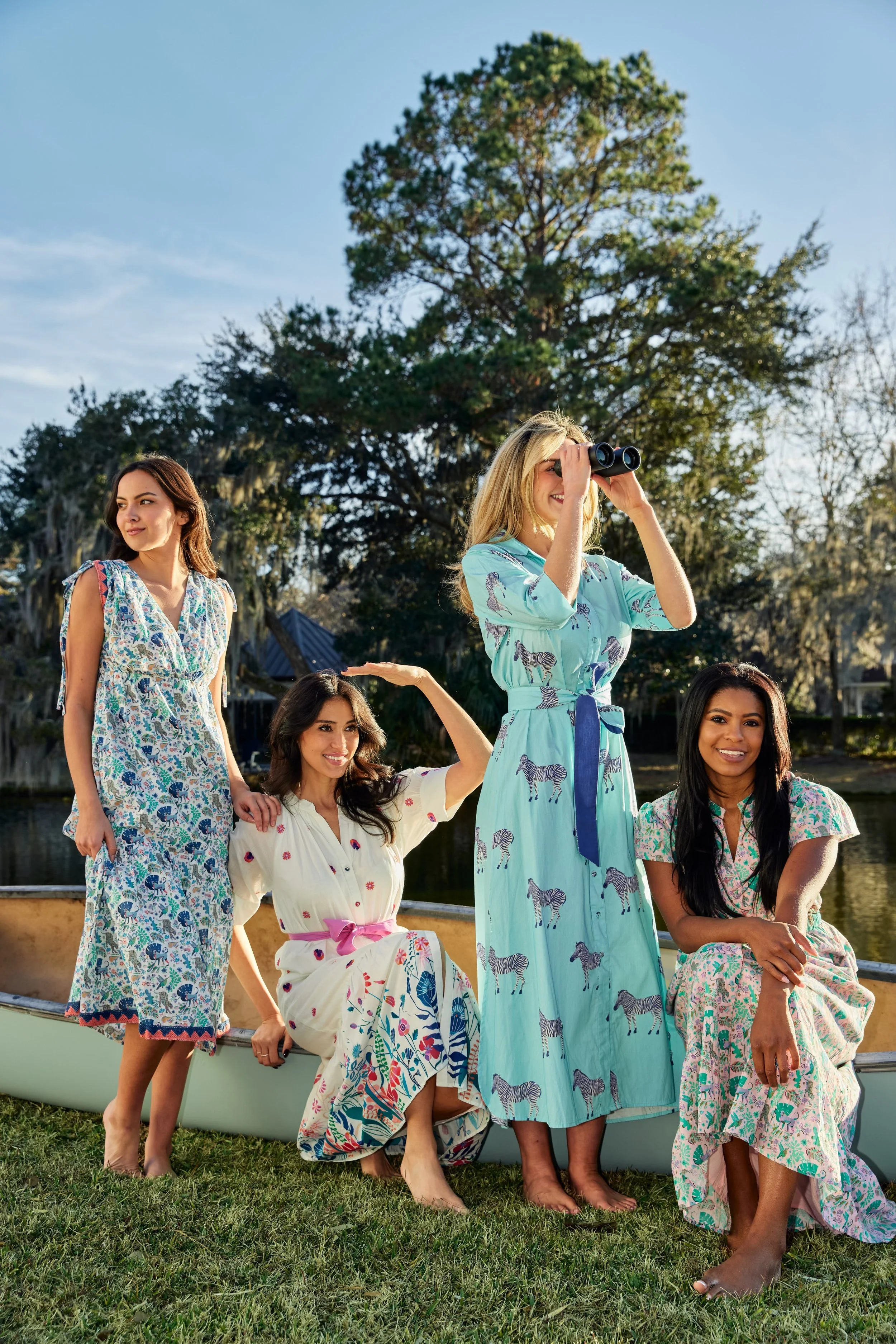 Group of women in colorful dresses on a boat near a lake, with trees and a house in the background under a clear sky.