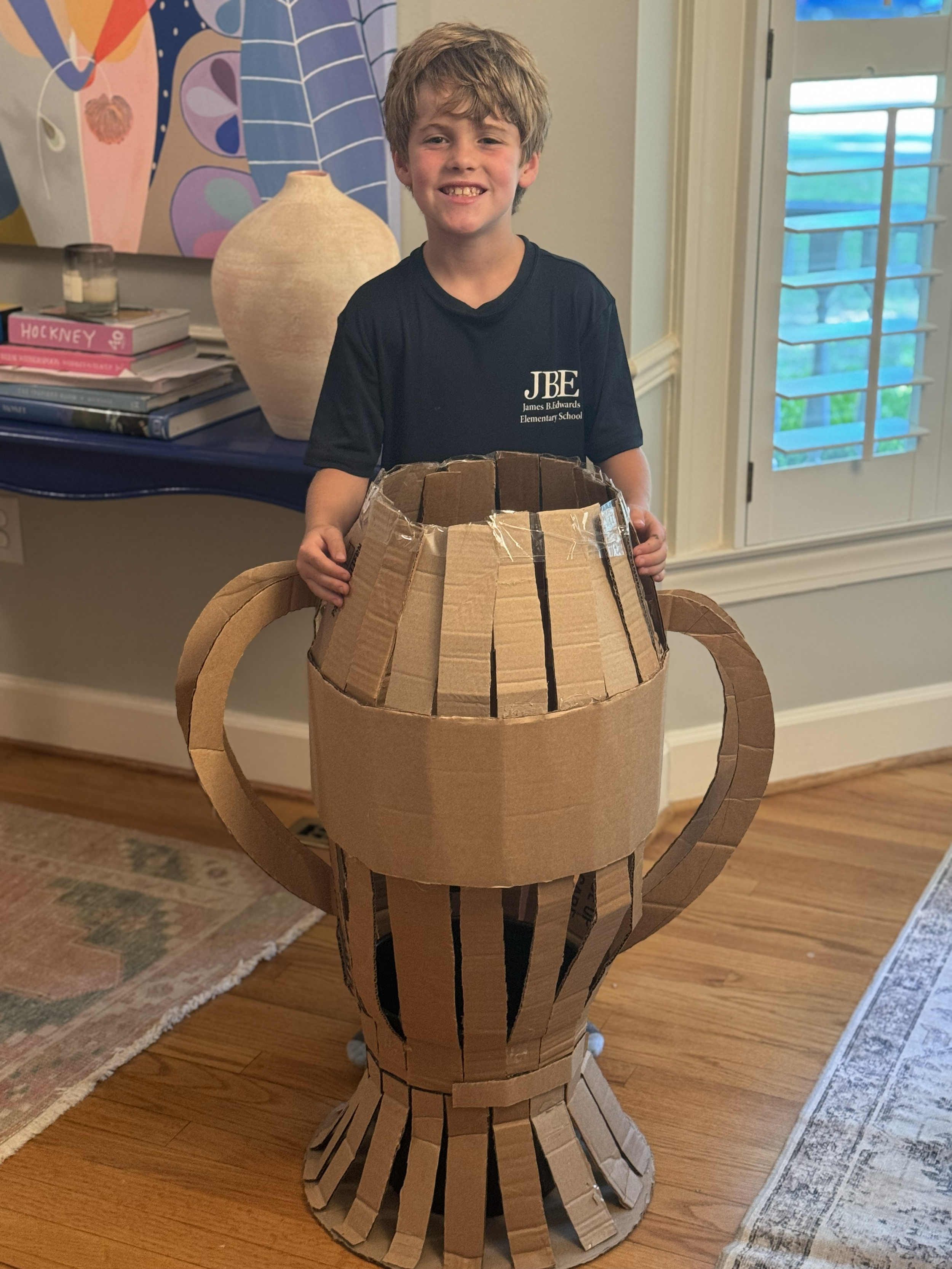 A young boy standing indoors, smiling, inside a large cardboard hot air balloon costume with a basket, made of recycled cardboard pieces, in a room with a blue table, stack of books, and a colorful abstract painting on the wall.