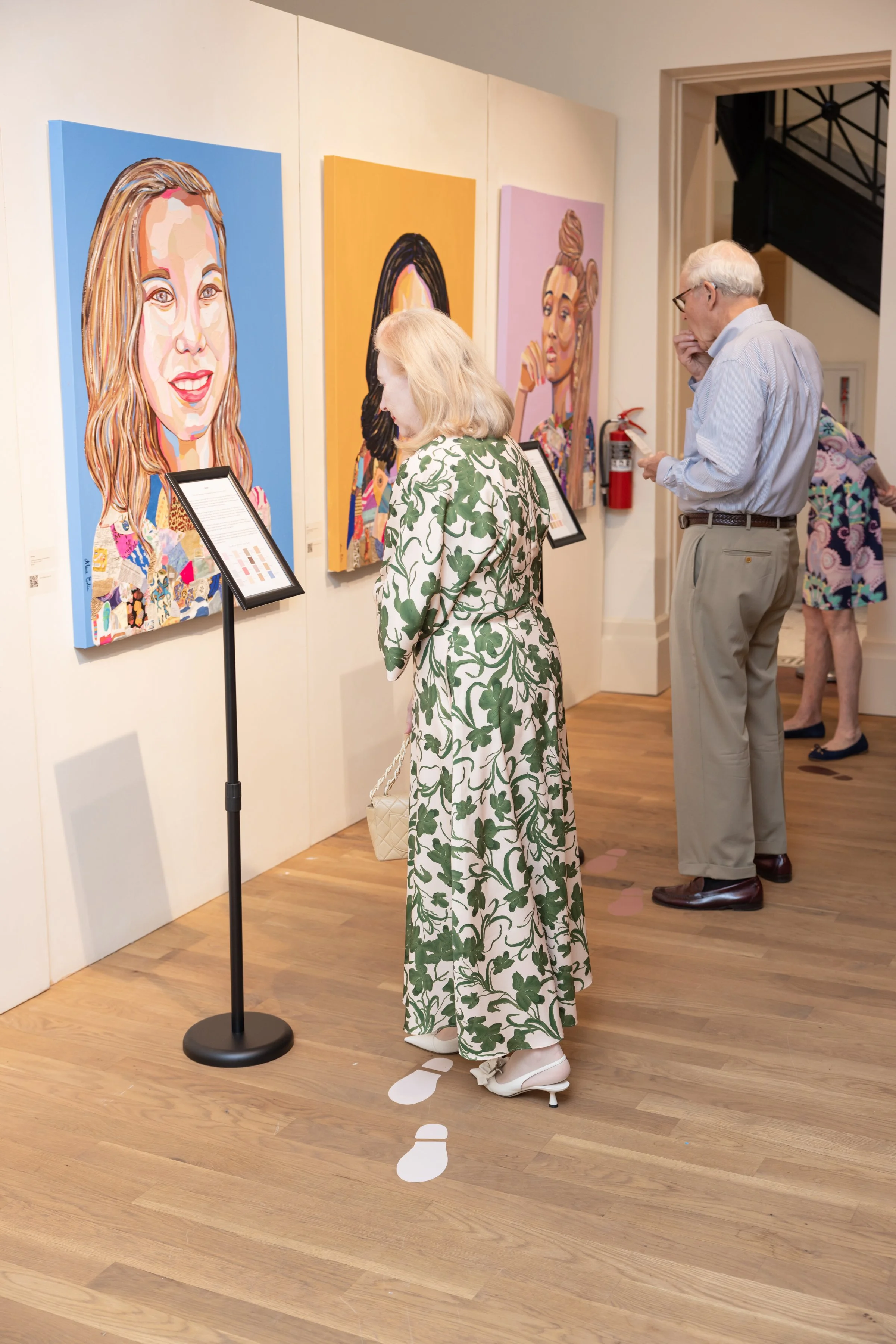 People viewing colorful portrait paintings in an art gallery. The woman in the foreground is dressed in a white dress with green leaf pattern and is wearing white shoes and standing on paper footprints on the wooden floor. The two men and a woman beh