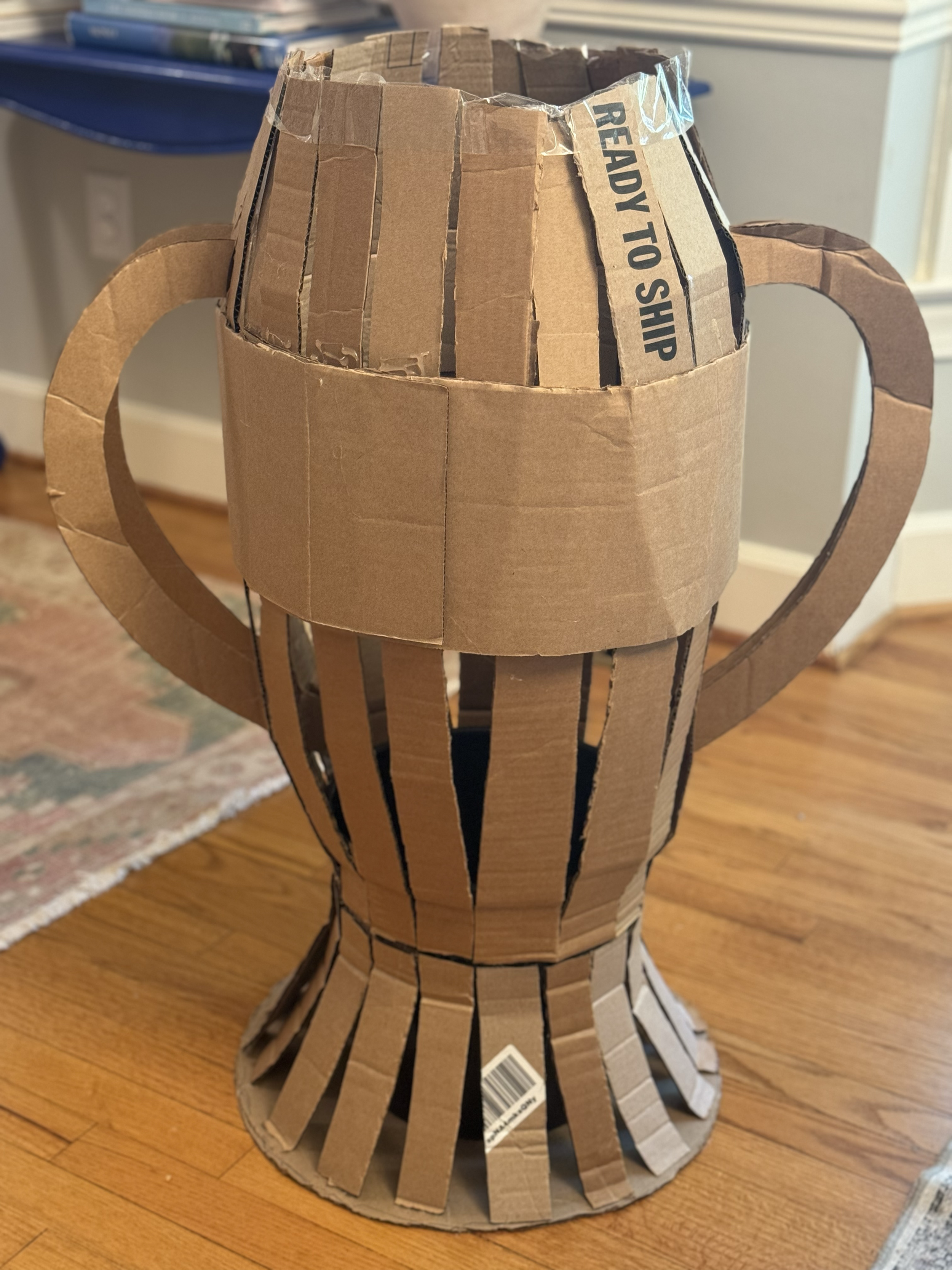 A cardboard sculpture of a person, resembling a trophy, made from brown cardboard and black tape, sitting on a hardwood floor.