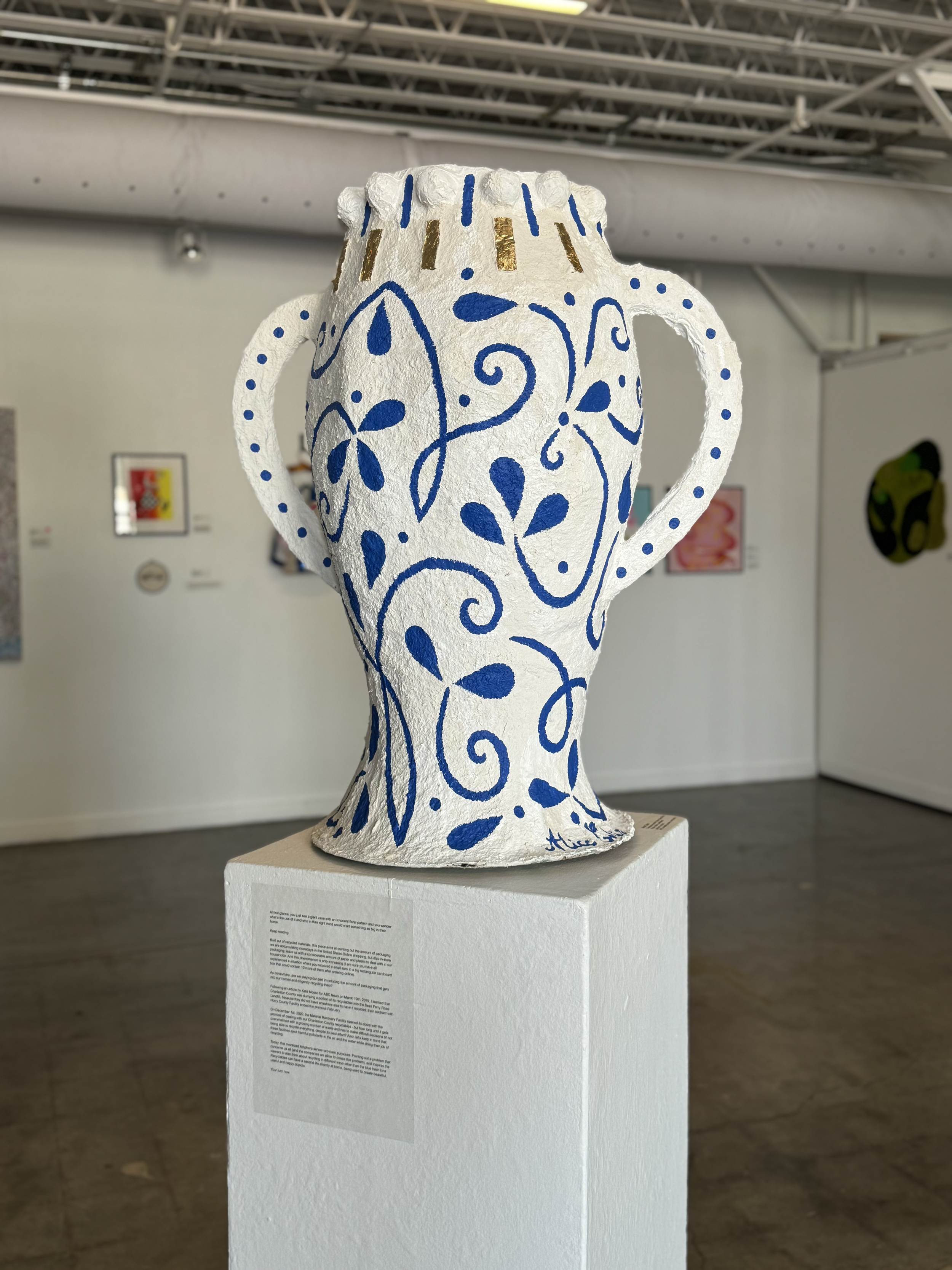 Ceramic vase sculpture painted with blue designs and gold accents, displayed on a white pedestal in an art gallery.