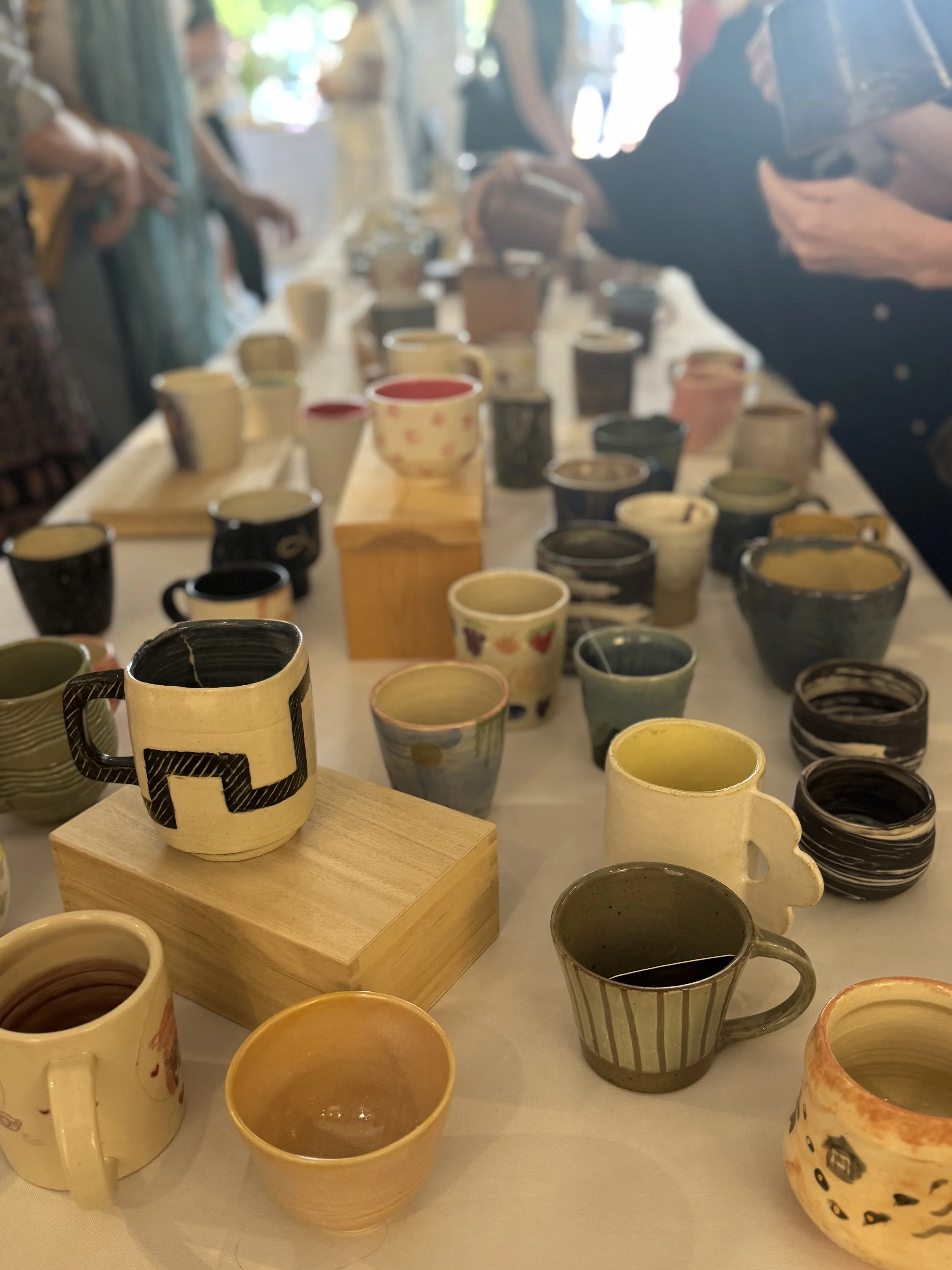 A table displaying various handmade ceramic mugs and cups with different colors, patterns, and shapes, some on wooden stands.
