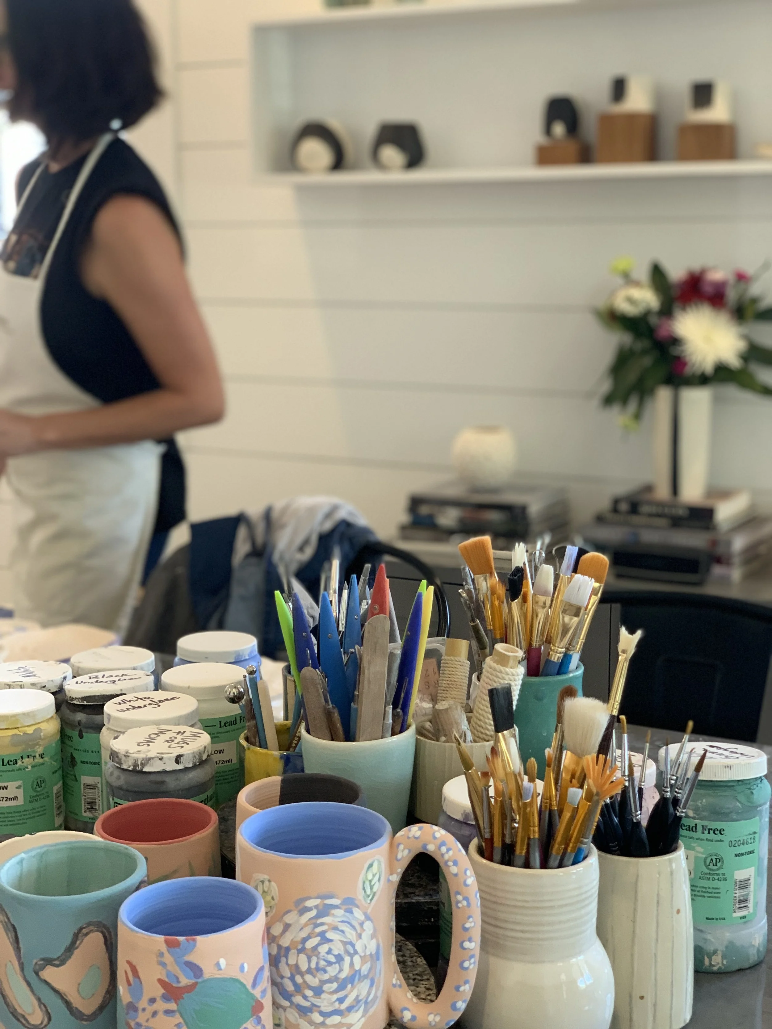 A table filled with various artist paint cups, brushes, and art supplies in the foreground. In the background, there is a woman with dark hair wearing a black top and cream apron, standing near a white wall with decorative black and wooden objects and a vase of flowers.