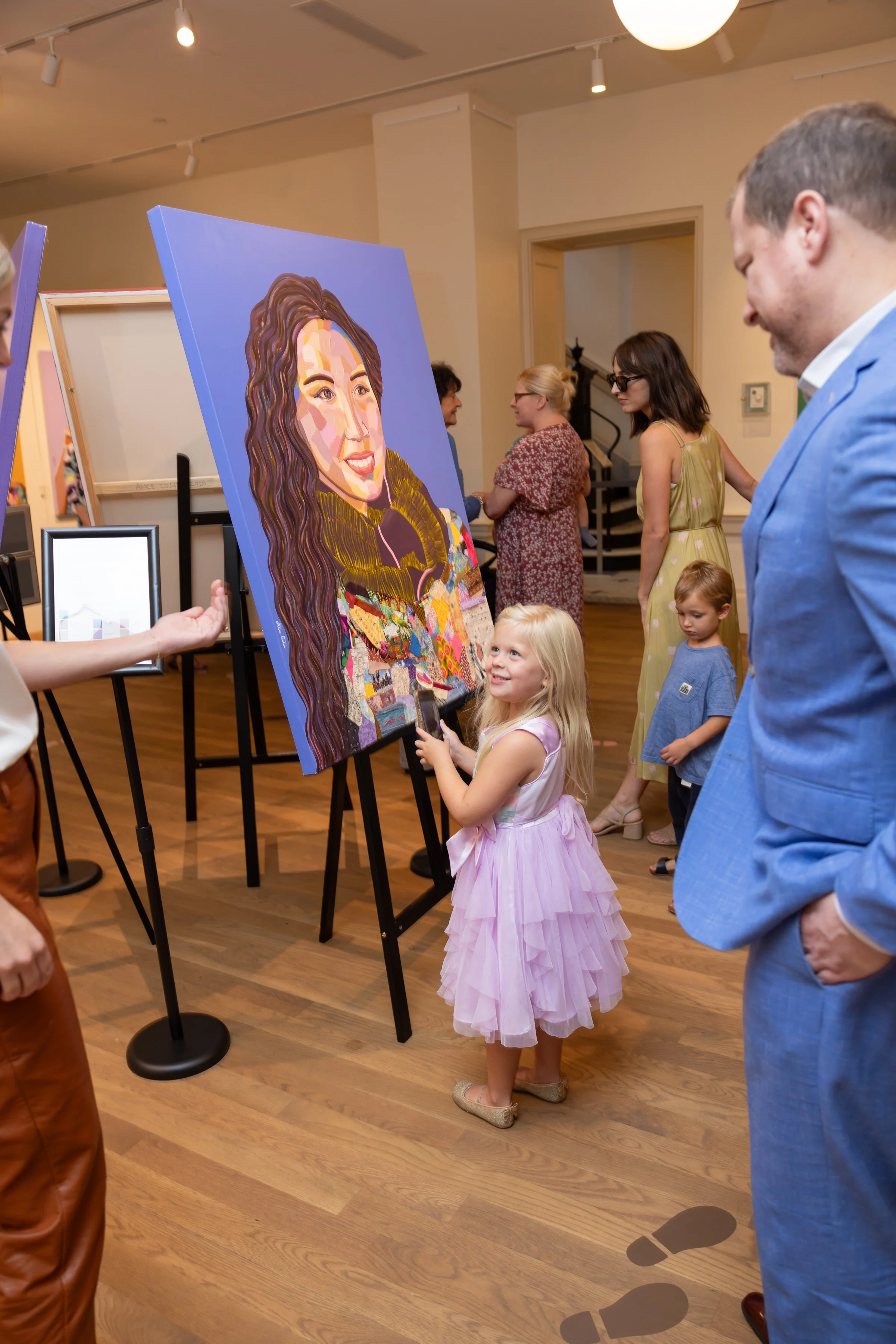 A girl in a pink dress looking at a colorful portrait painting of a woman with long wavy hair at an art gallery.