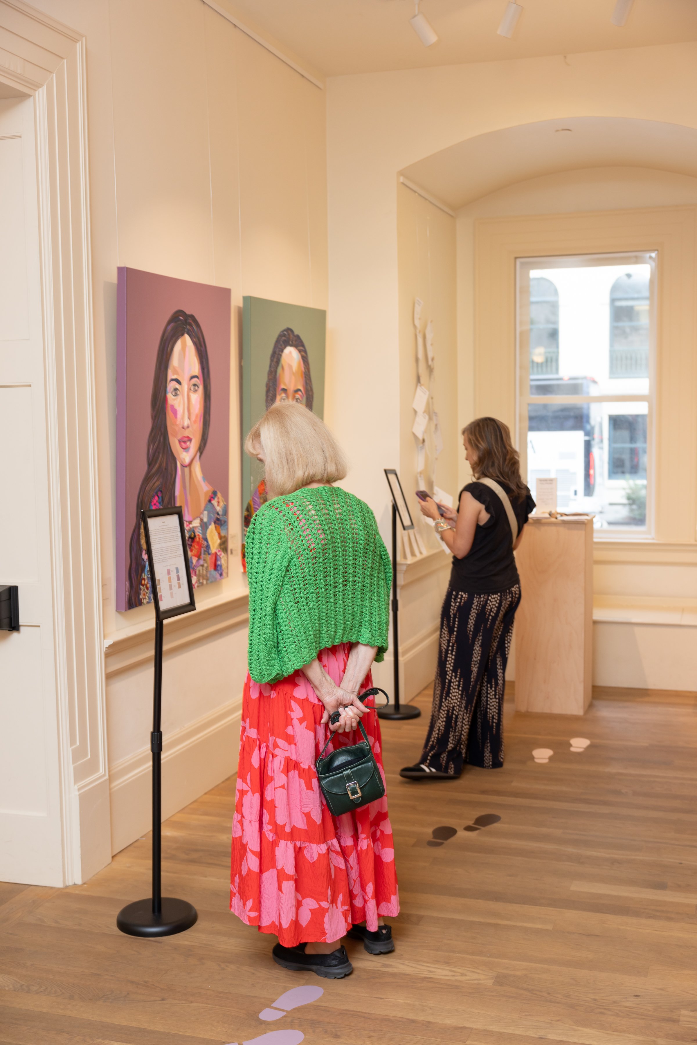 Three women stand inside an art gallery, viewing colorful portraits of women on the wall. One woman wears a pink floral skirt and a green crocheted jacket, holding a black handbag. The other woman is dressed in black patterned pants and a black top, 