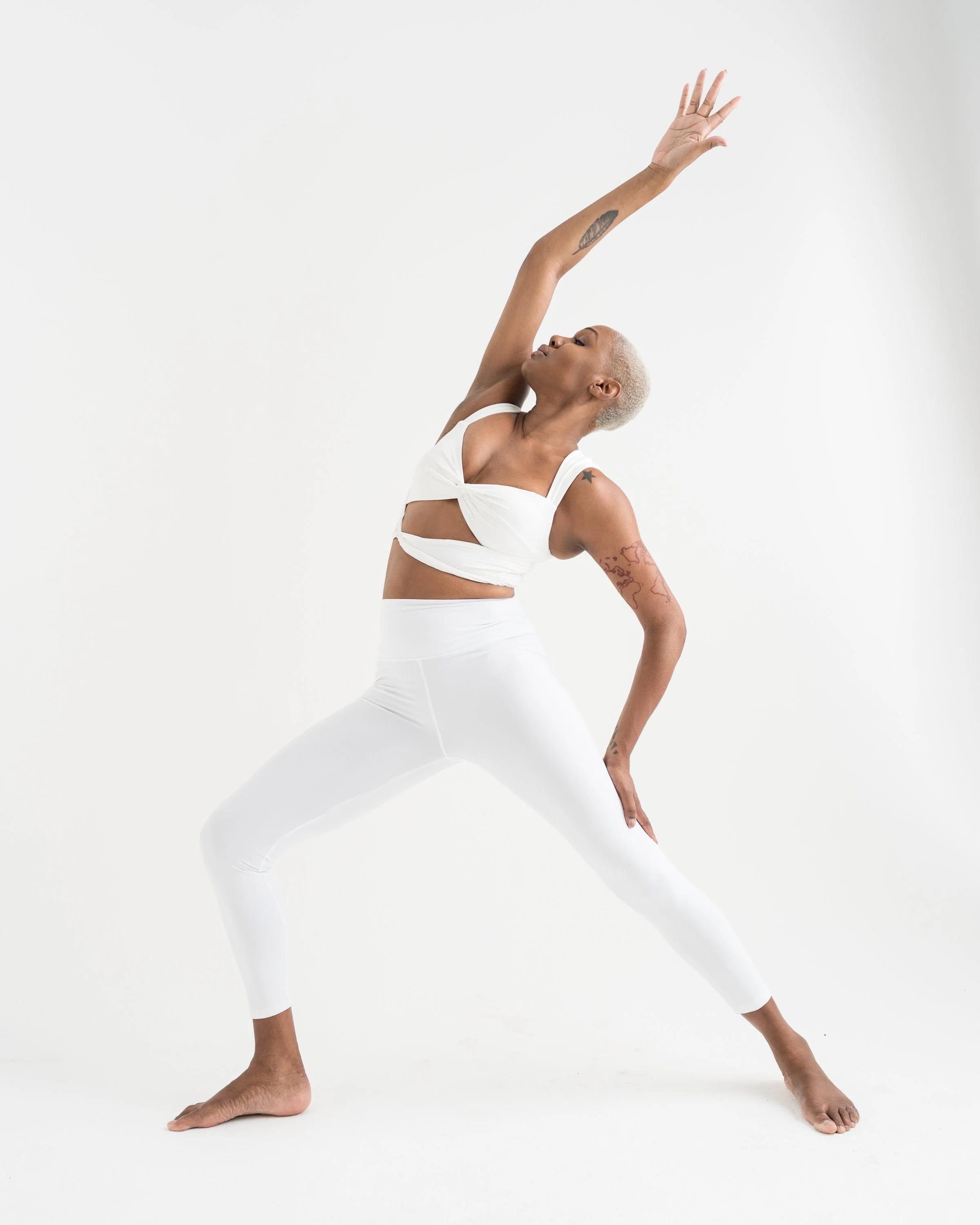 A woman with light blonde hair and tattoos on her arms is performing a yoga pose against a white background, wearing a white workout outfit.