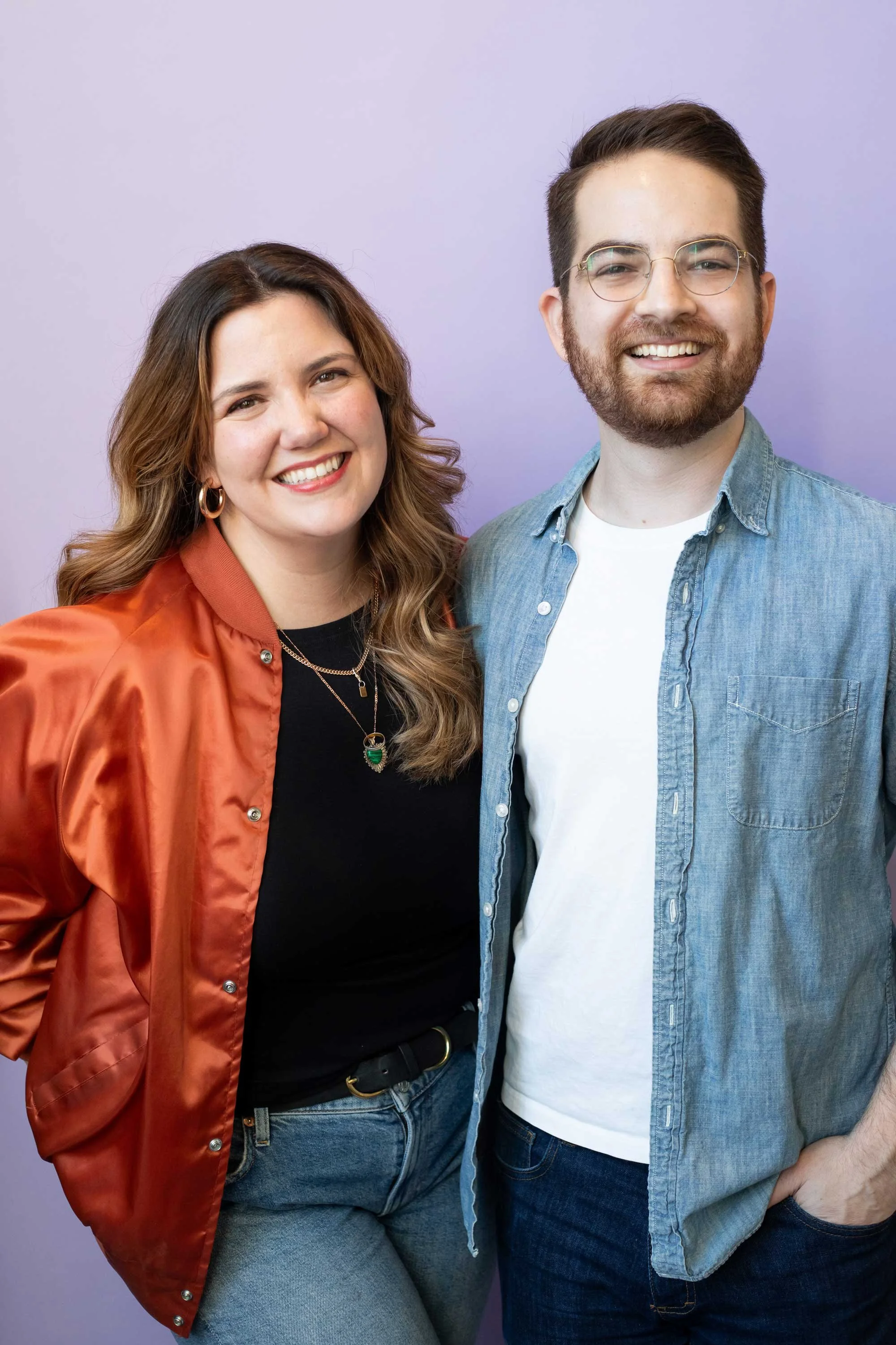 A smiling woman with wavy brown hair wearing a black shirt, orange satin jacket, and jeans standing next to a smiling man with short dark hair, glasses, a beard, wearing a light blue denim shirt and a white T-shirt, against a light purple background.