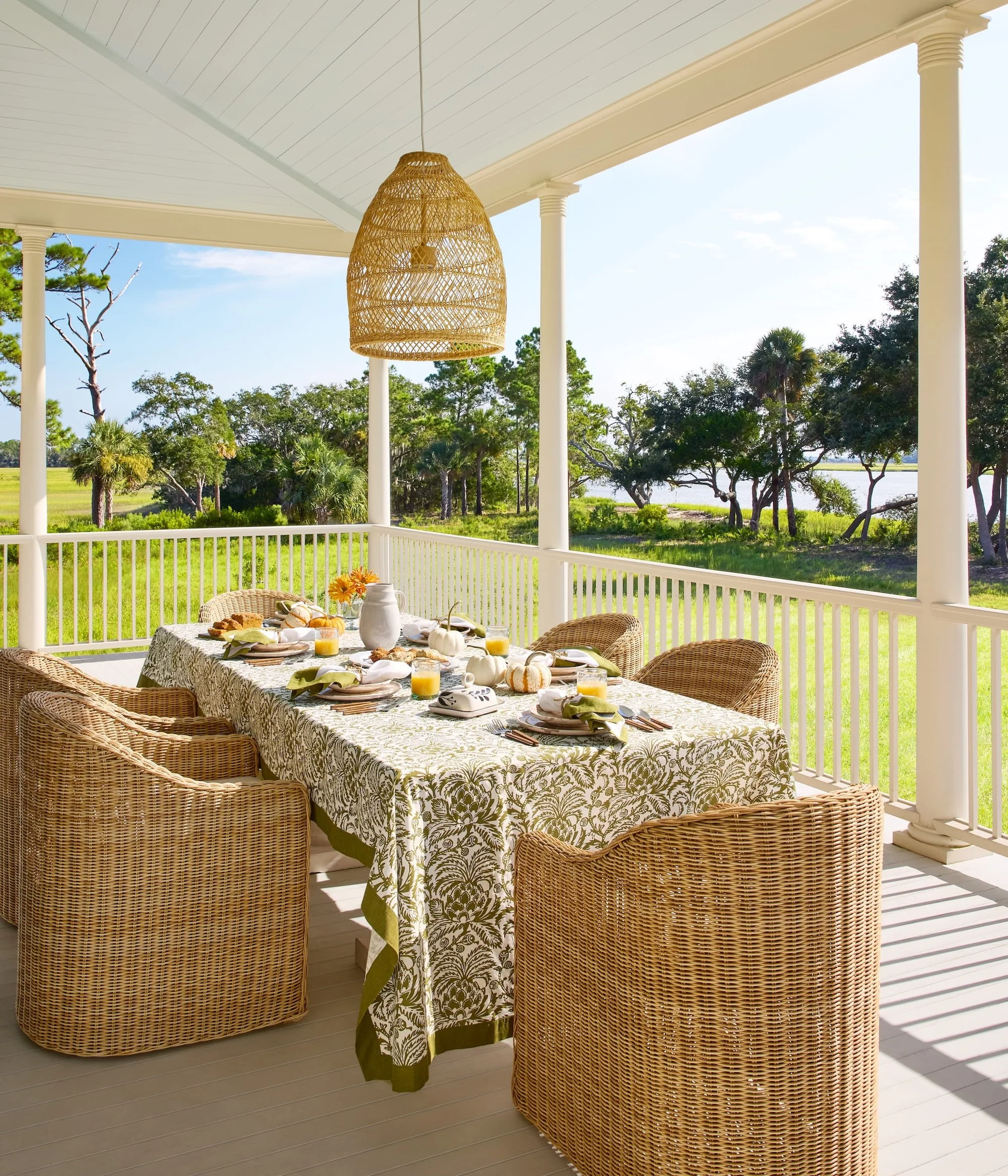 A porch with a white railing overlooking a lush green landscape with trees and water in the distance. There is a dining table covered with a patterned tablecloth, set with dishes, glasses, and a white vase with orange flowers. wicker chairs surround 
