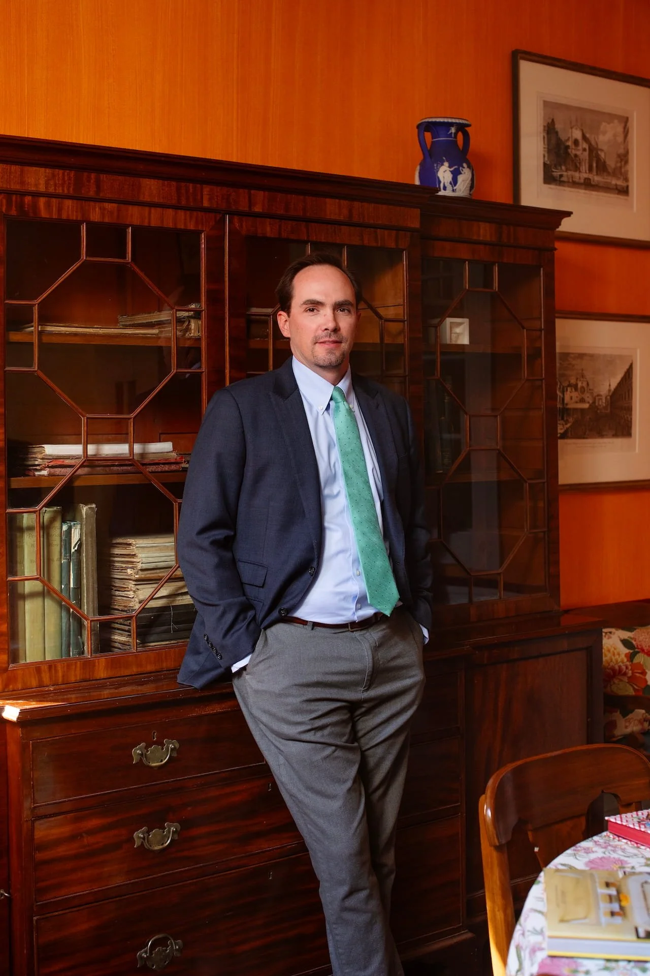 A man in a suit with a green tie stands in front of a wooden cabinet in a room with wood-paneled walls, framed pictures, and a decorative blue vase on top of the cabinet.