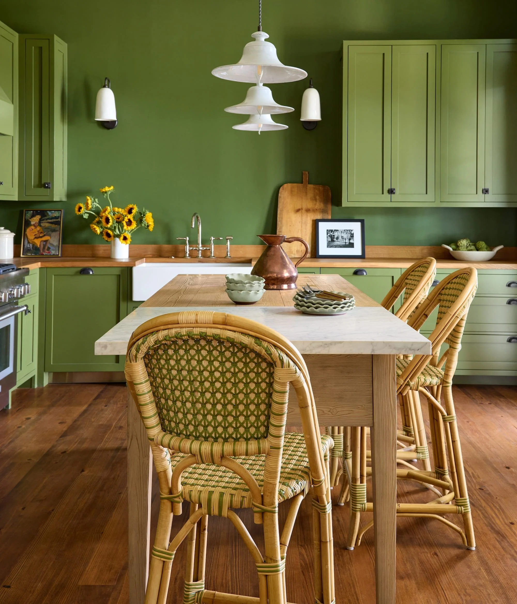 Kitchen with green cabinets, wooden countertop, hanging white pendant lights, sunflower bouquet, framed artwork, and rattan chairs around a marble-topped table.
