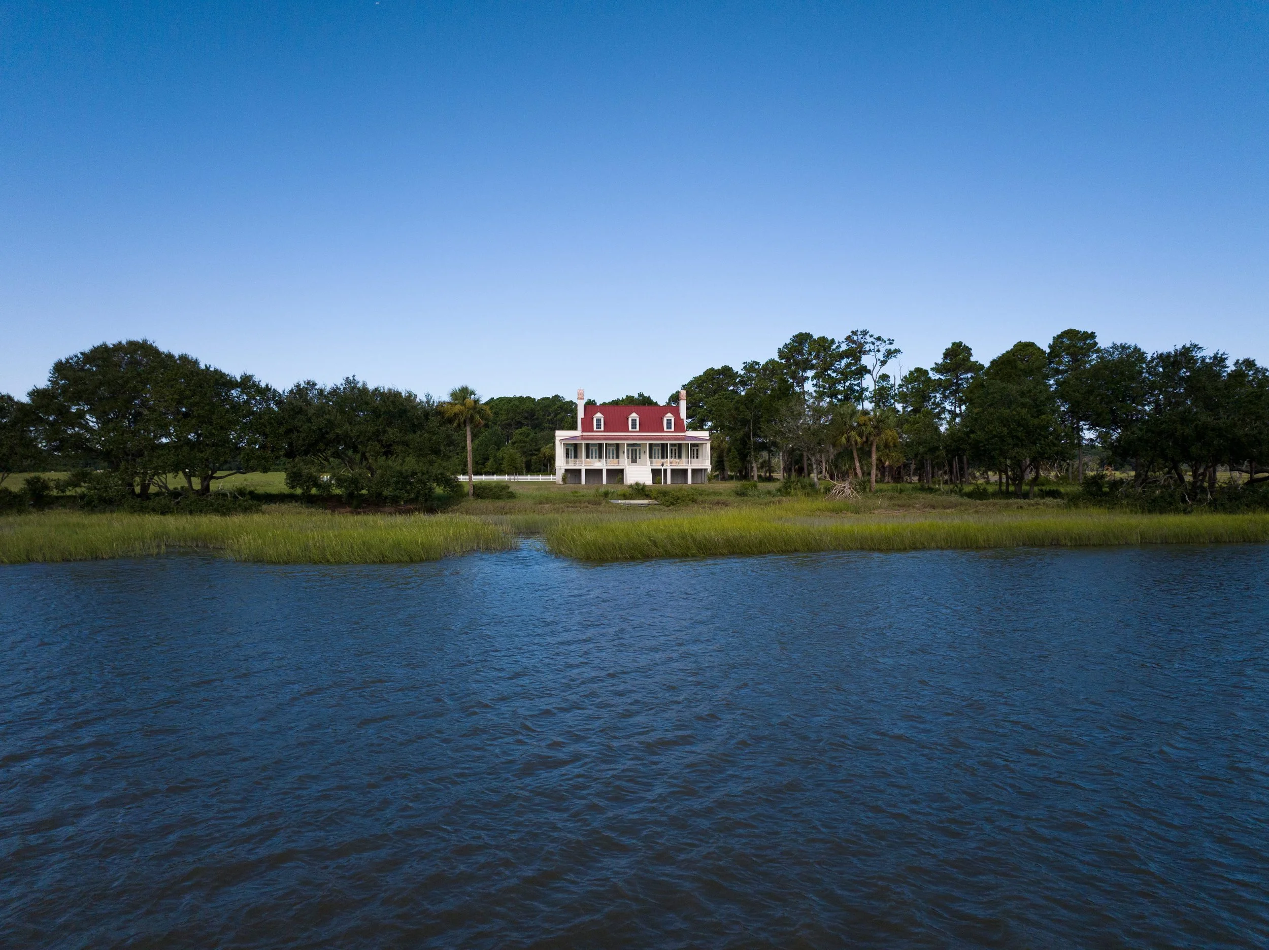 A house with a red roof sitting on the edge of a water body, surrounded by trees and greenery, under a clear blue sky.