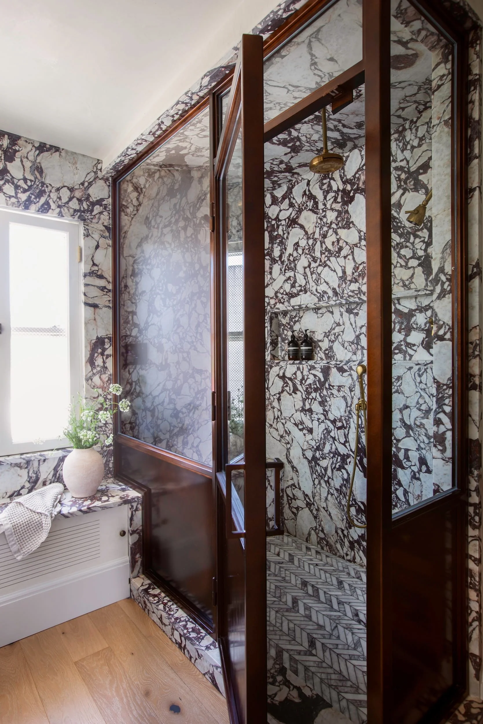 A bathroom with marble walls, a walk-in shower with a glass door, metallic fixtures, and a window with a plant on a nearby cabinet.