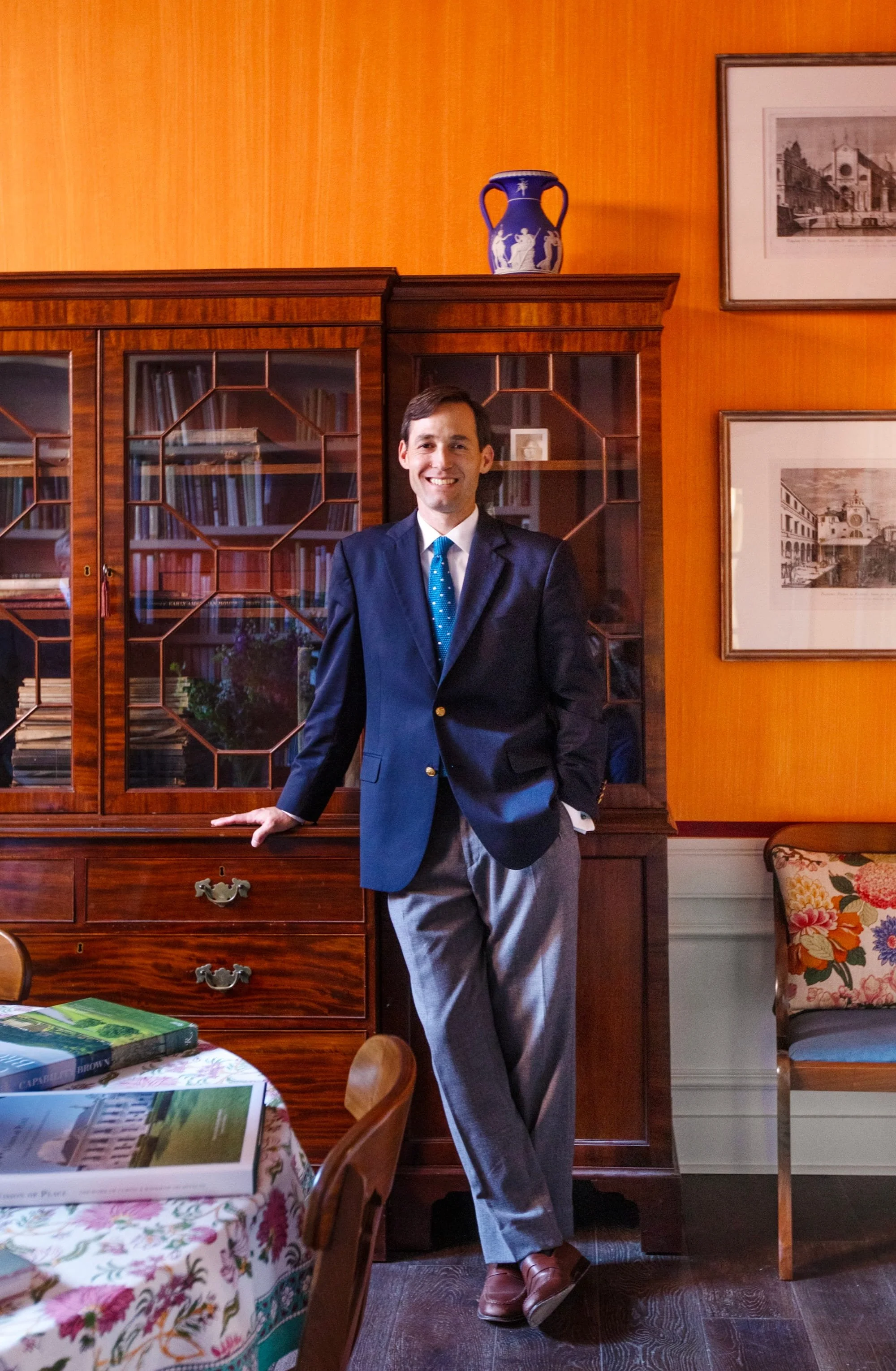 A man dressed in a navy blazer, white shirt, and gray trousers, standing in a room with orange walls, a wooden cabinet, and framed artwork. He is smiling with his left hand resting on the cabinet.