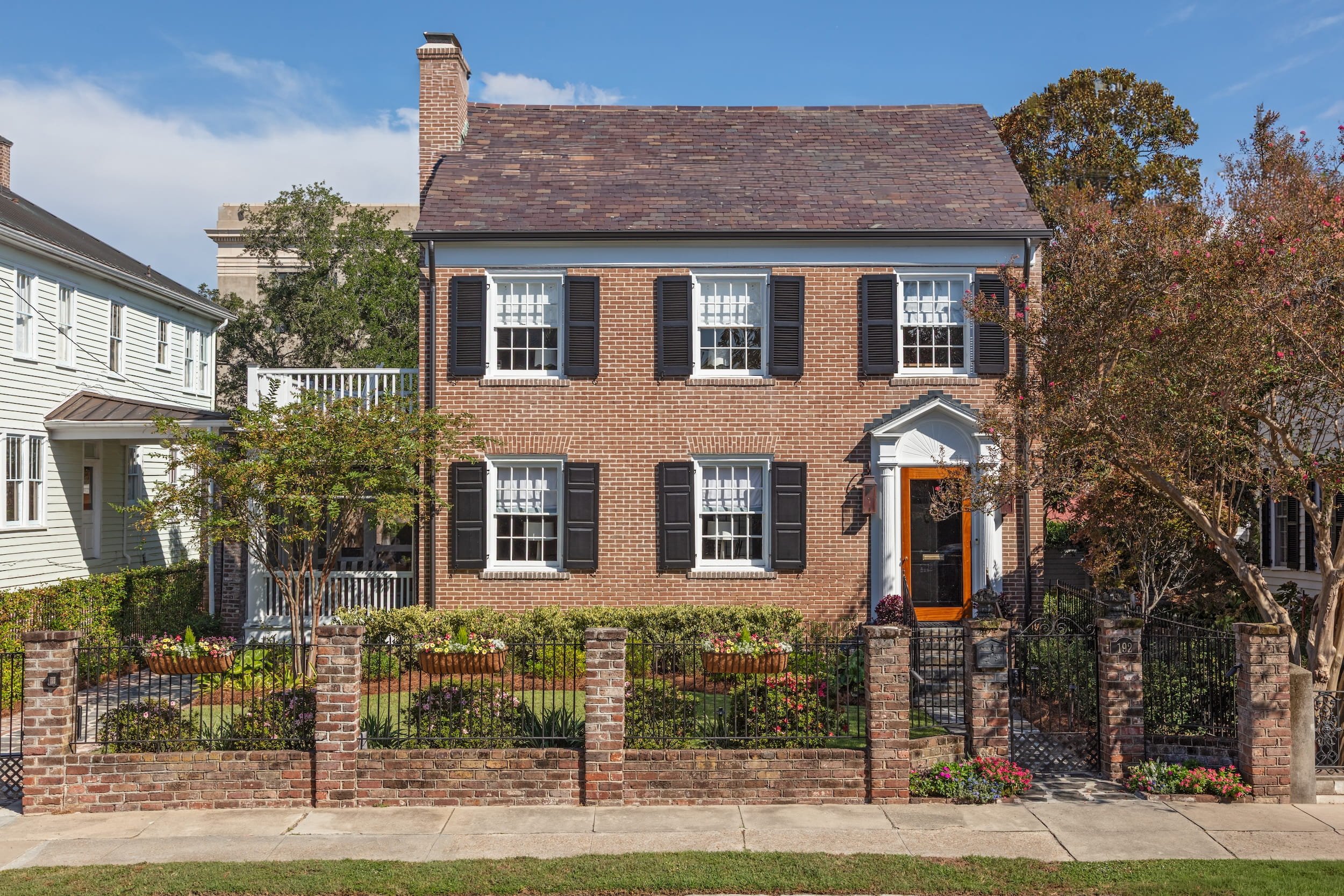 A brick house with black shutters, white window frames, a black front door with white decorative trim, and a small front yard enclosed by a brick and black metal fence.
