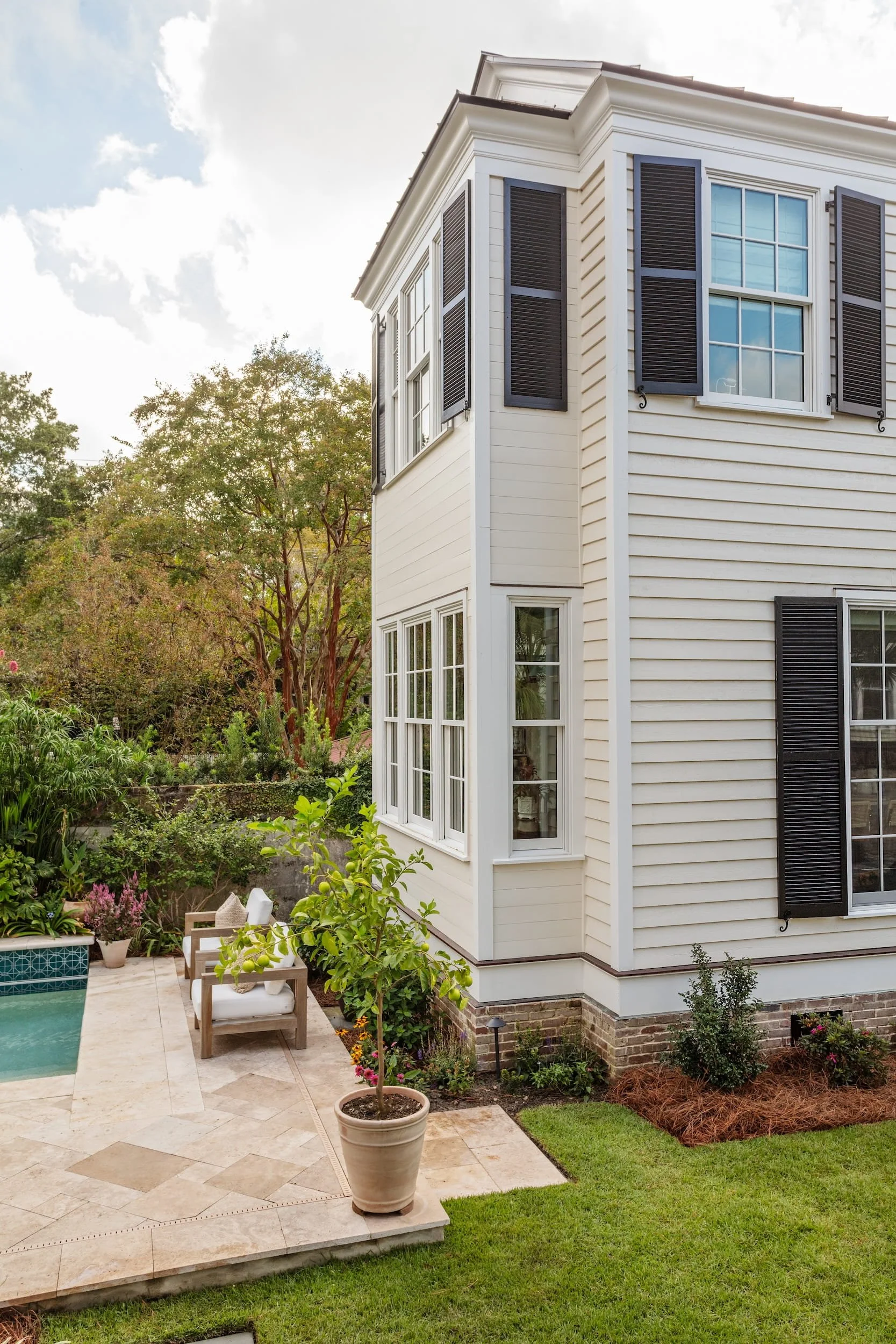 View of the backyard patio area of a three-story house with beige siding, black shutters, and numerous windows. The patio has a small table with chairs, a potted plant, and garden beds with plants and flowers. There is a garden with trees and shrubs 