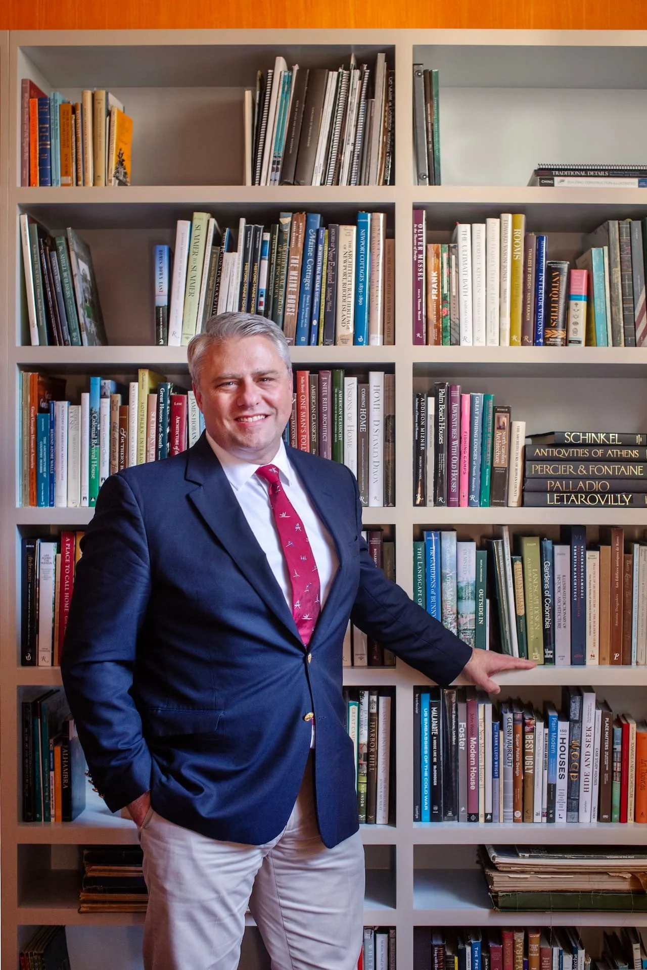 A smiling man in a dark blazer, white shirt, and red tie standing in front of a bookshelf filled with various books, with his right hand pointing to books on the shelf.