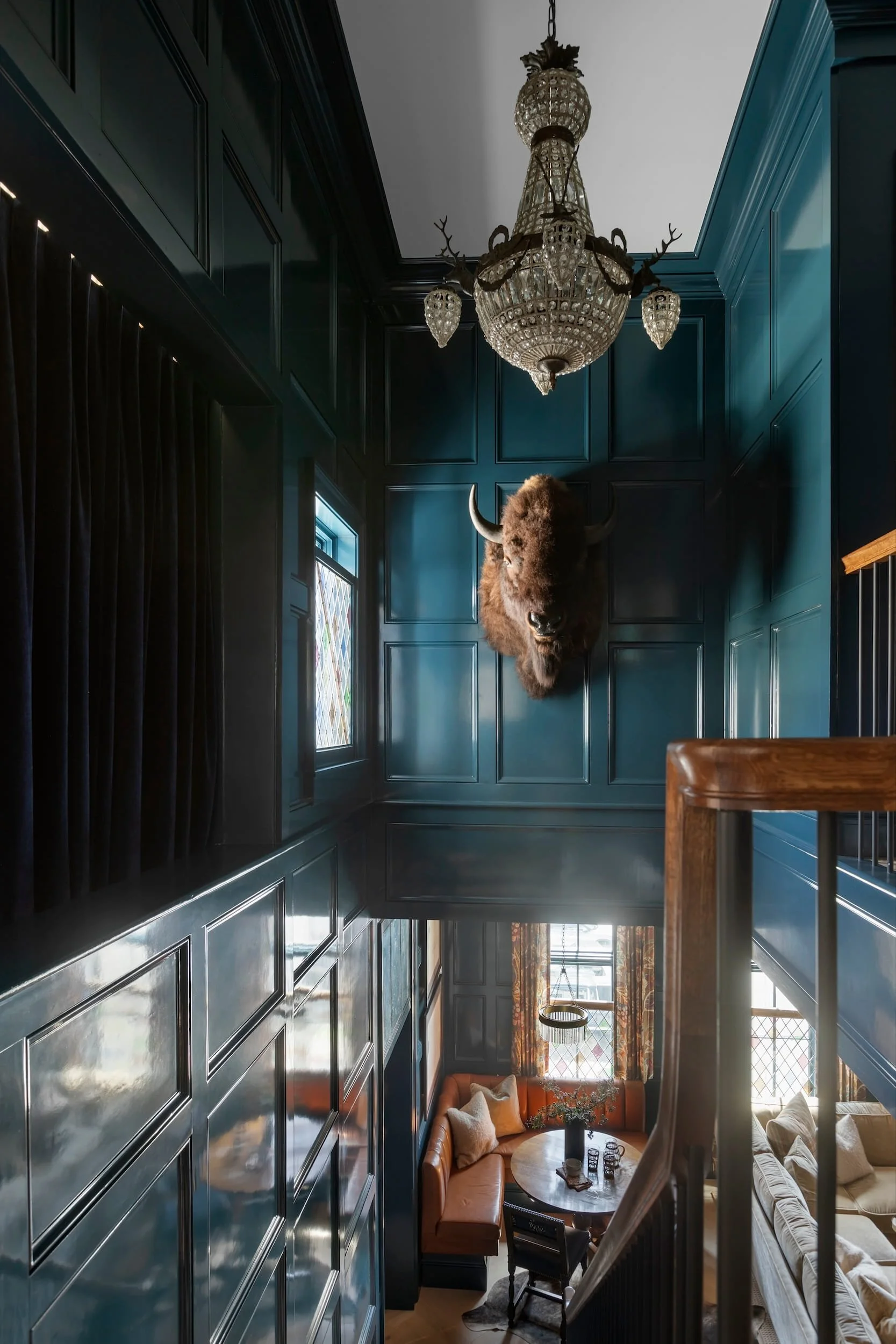 Interior view of a home with high dark paneled walls, a large ornate chandelier hanging from the ceiling, a mounted buffalo head with horns, stained glass windows, and cozy seating area with orange and beige sofas and a round coffee table.