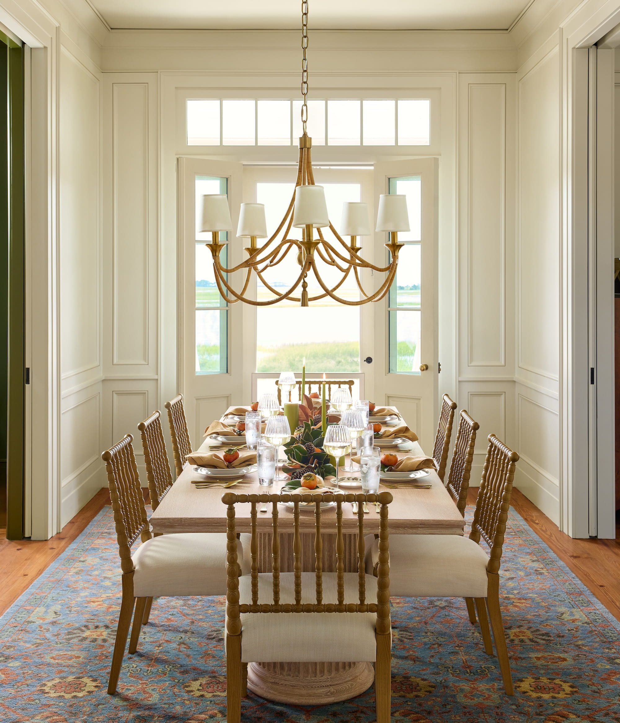 Dining room with a long table set for a meal, decorated with candles and greenery, surrounded by wooden chairs, in front of large windows with a view of fields and sky, and a brass chandelier hanging above.