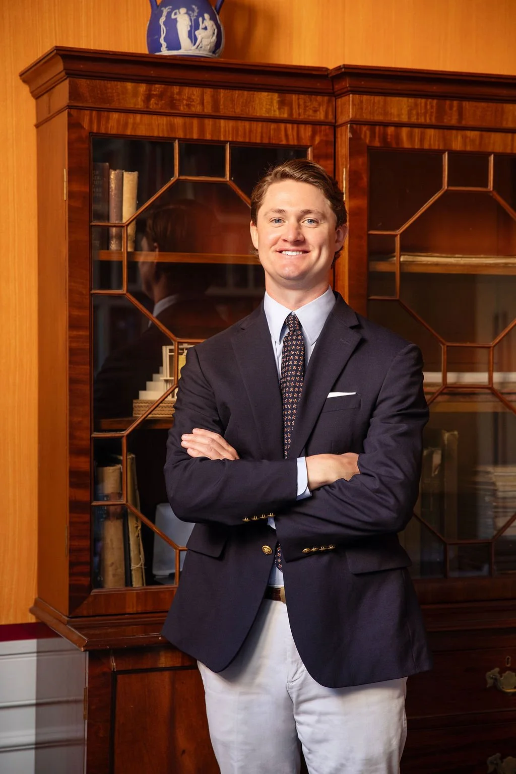 A man in coat and tie standing in front of a breakfront with his arms crossed over his chest, smiling at the camera.