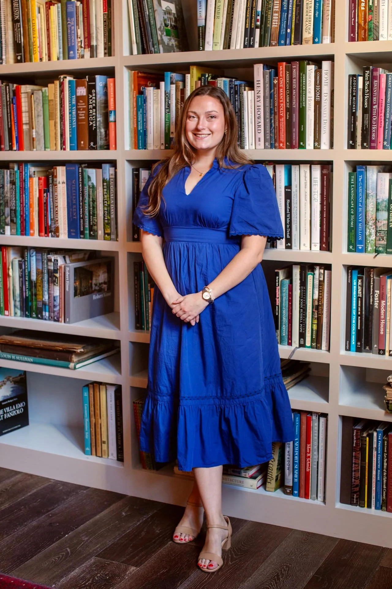 A woman with long brown hair wearing a blue dress with puffed sleeves stands in front of a bookshelf filled with numerous books, smiling at the camera.