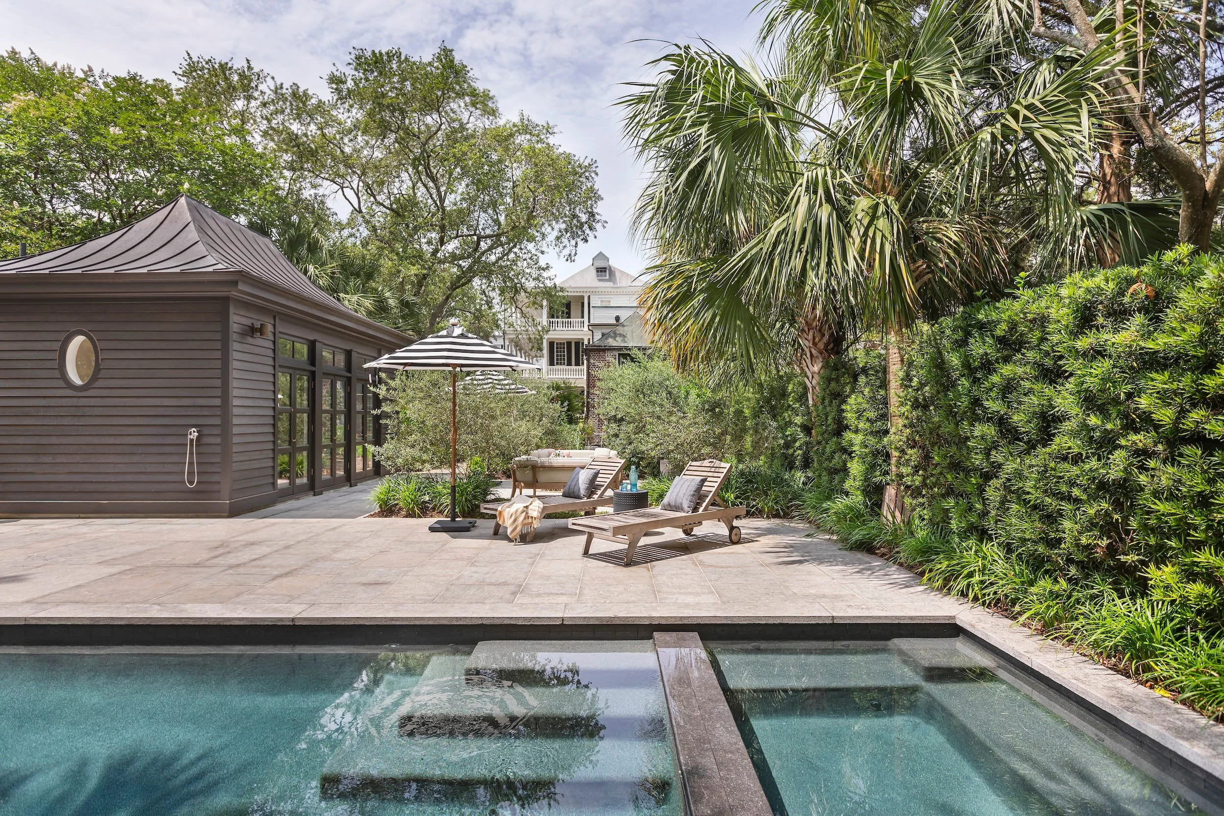 A backyard patio with lounge chairs, striped umbrella, outdoor sofa, and a swimming pool, surrounded by green trees and shrubs, with a house in the background.