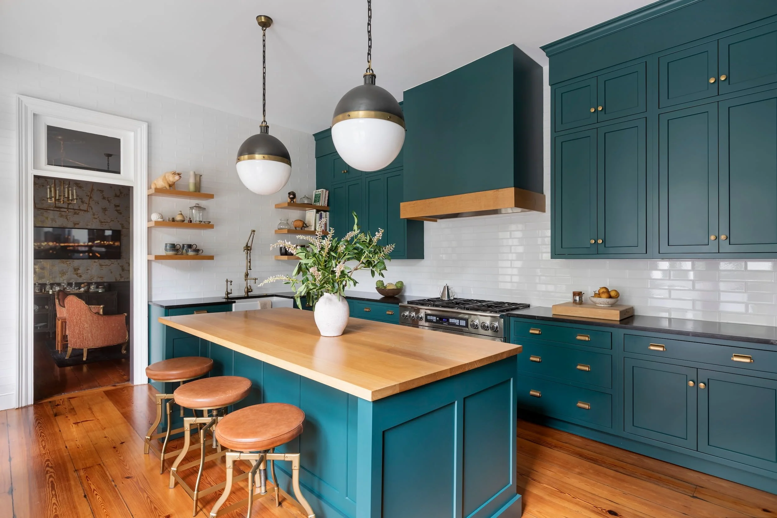 Modern kitchen with teal cabinets, a wooden island, and pendant lights. Includes a white brick backsplash, open shelves with decorative items, and a vase with flowers on the island.