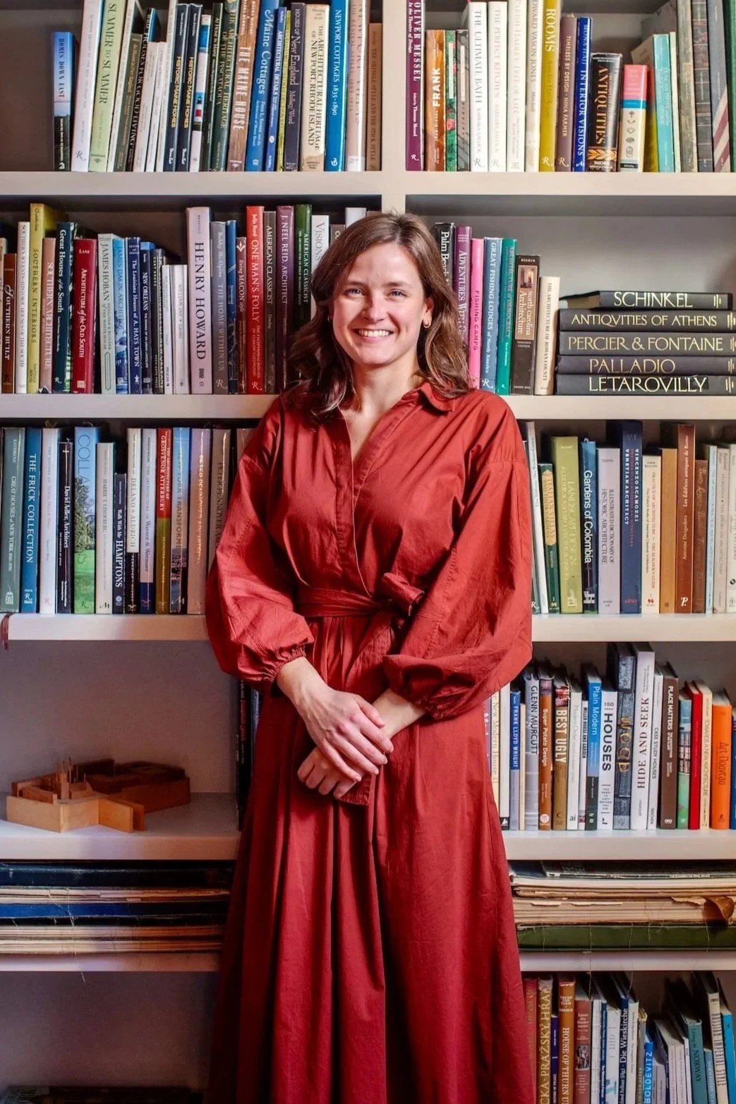 A woman standing in front of a bookshelf filled with various books. She is wearing a long, red dress with elasticized sleeves and is smiling at the camera.