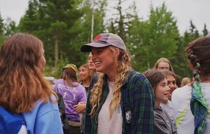 Group of people, mostly young women, outdoors in a forested area, smiling and engaging in conversation.
