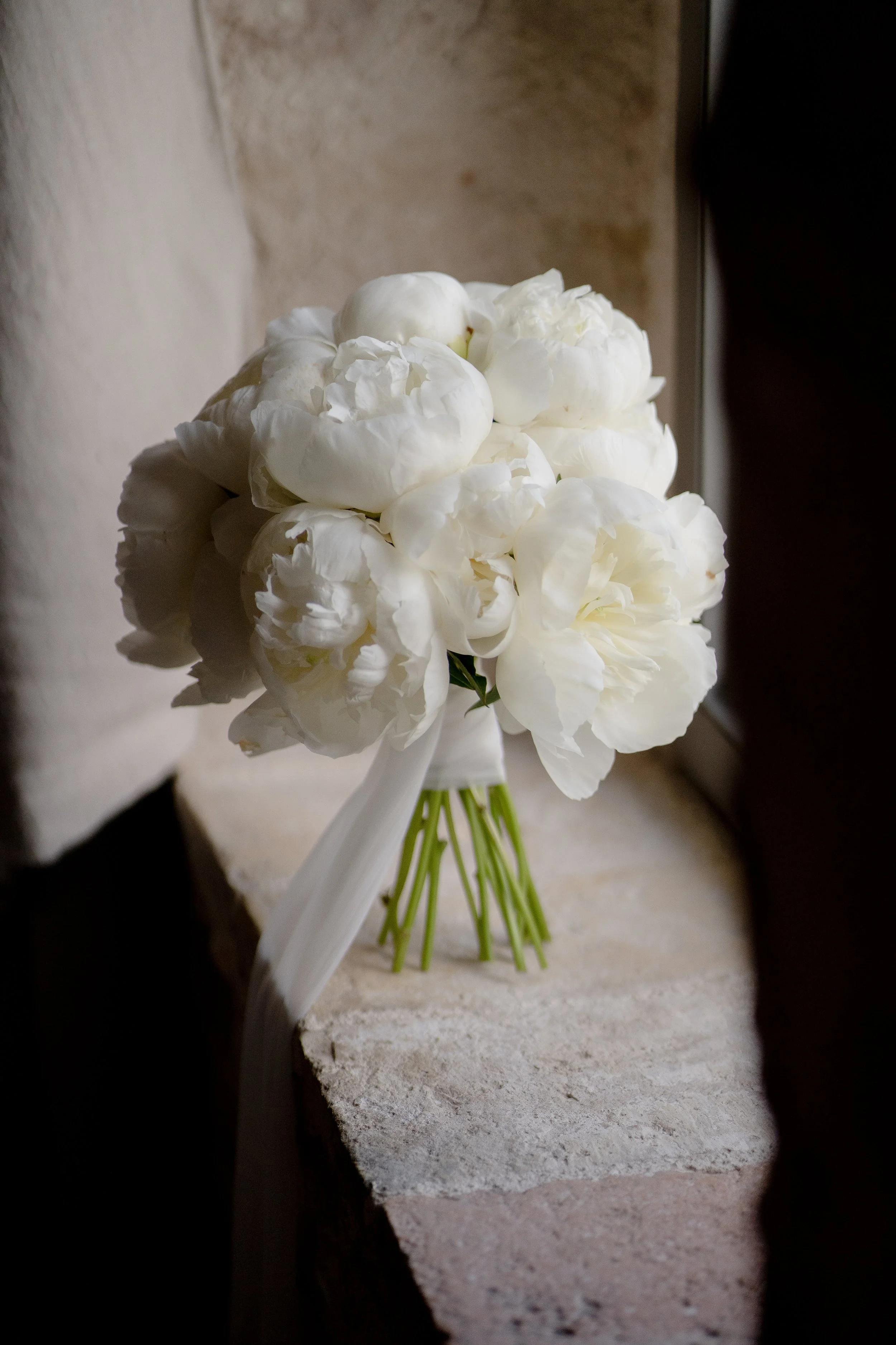 A bouquet of white flowers with green stems resting on a ledge with a plain wall in the background.