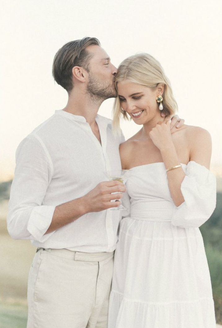 A man kisses a woman on the head while holding a glass, both dressed in white, outdoors during daylight, smiling and enjoying a moment together.
