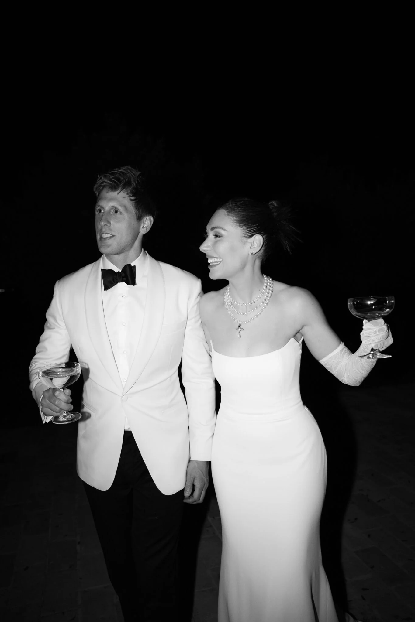 A man and woman dressed in formal wedding attire at night, holding champagne glasses. The man wears a white tuxedo with a black bow tie, and the woman wears a white strapless wedding gown with pearl necklaces and lace gloves. They are smiling and looking to the side.