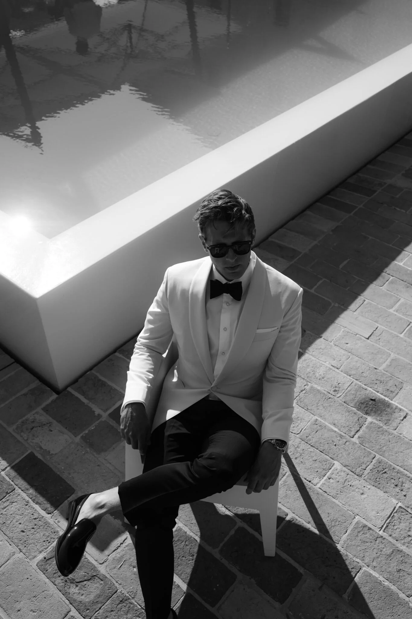 A man in a white tuxedo and black bow tie sitting on a white chair beside a swimming pool, wearing sunglasses and black dress shoes, with shadows cast on the brick ground.