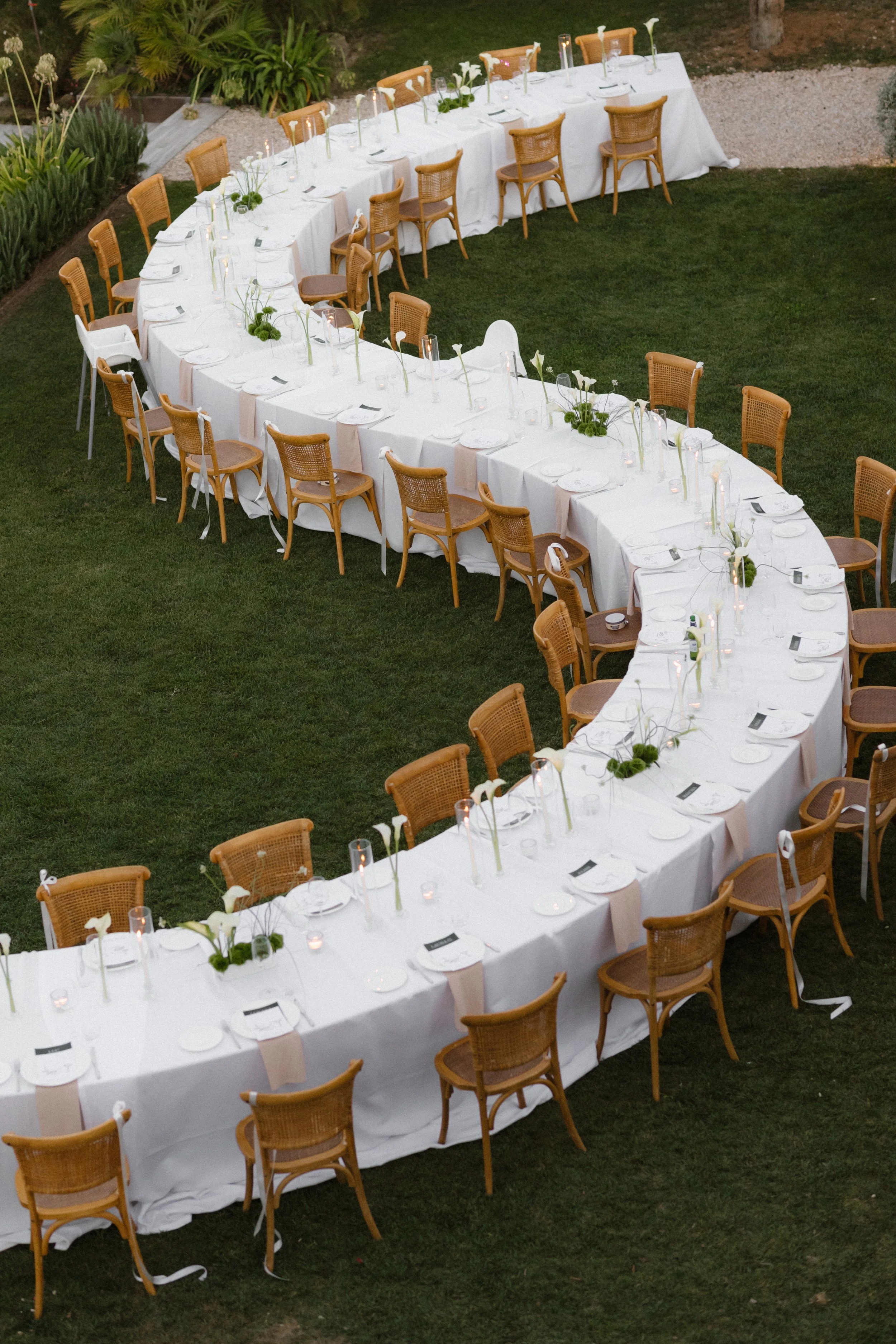 Long banquet table set for a formal outdoor event, covered with white tablecloths, small floral centerpieces, tall candles, and arranged with chairs on both sides, on a grassy area with plants in the background.