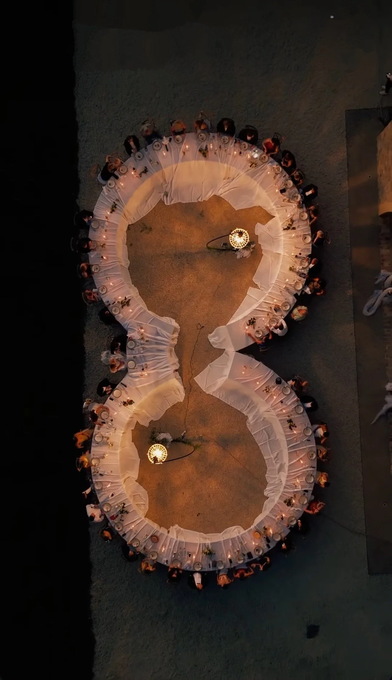 Overhead view of a large round dinner table at a social event, with many people seated around it, lit by hanging lamps. The table is covered with a white tablecloth, plates, and floral centerpieces, on a gravel or concrete surface.
