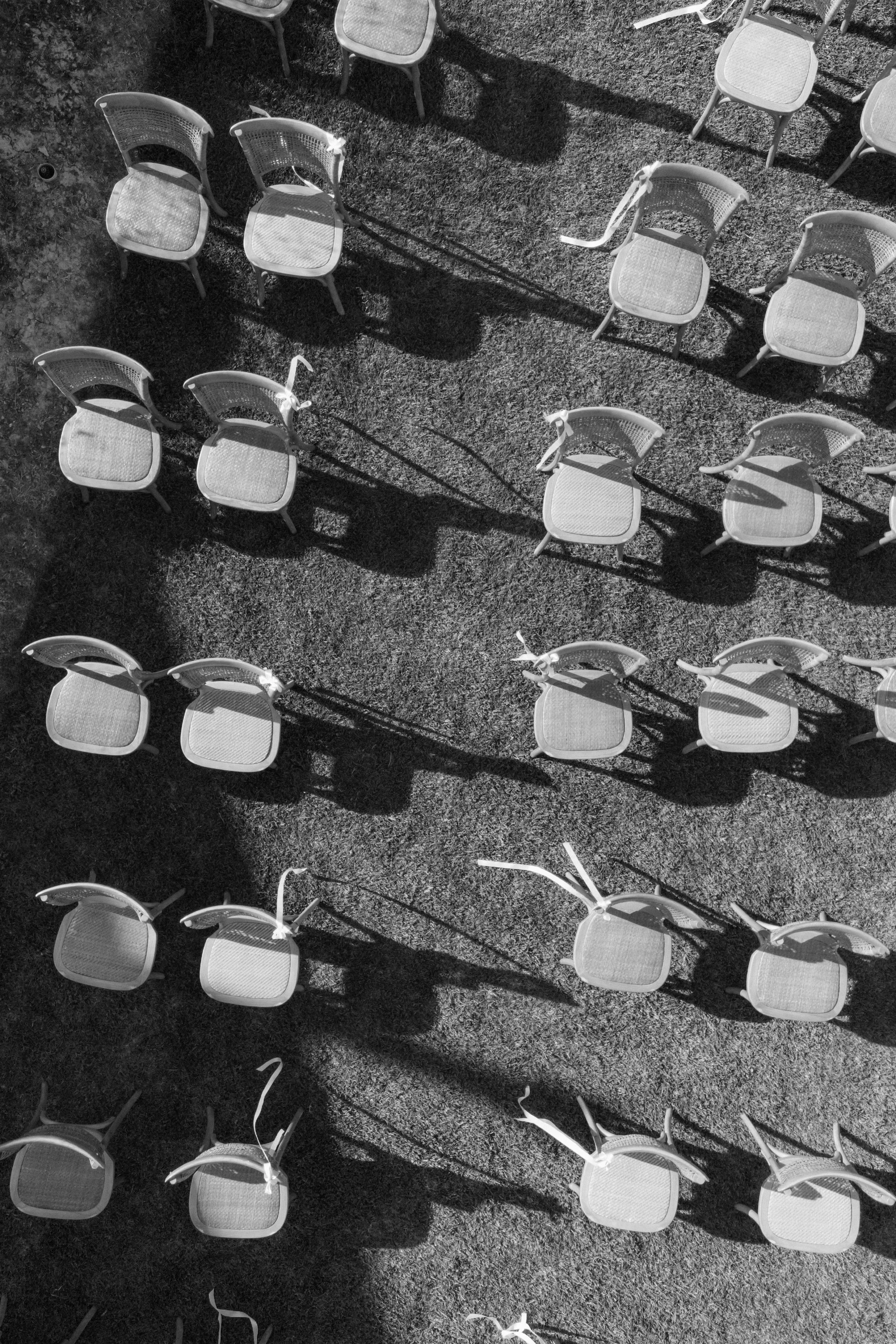 Black and white aerial view of chairs arranged on grass, with their shadows cast on the ground.