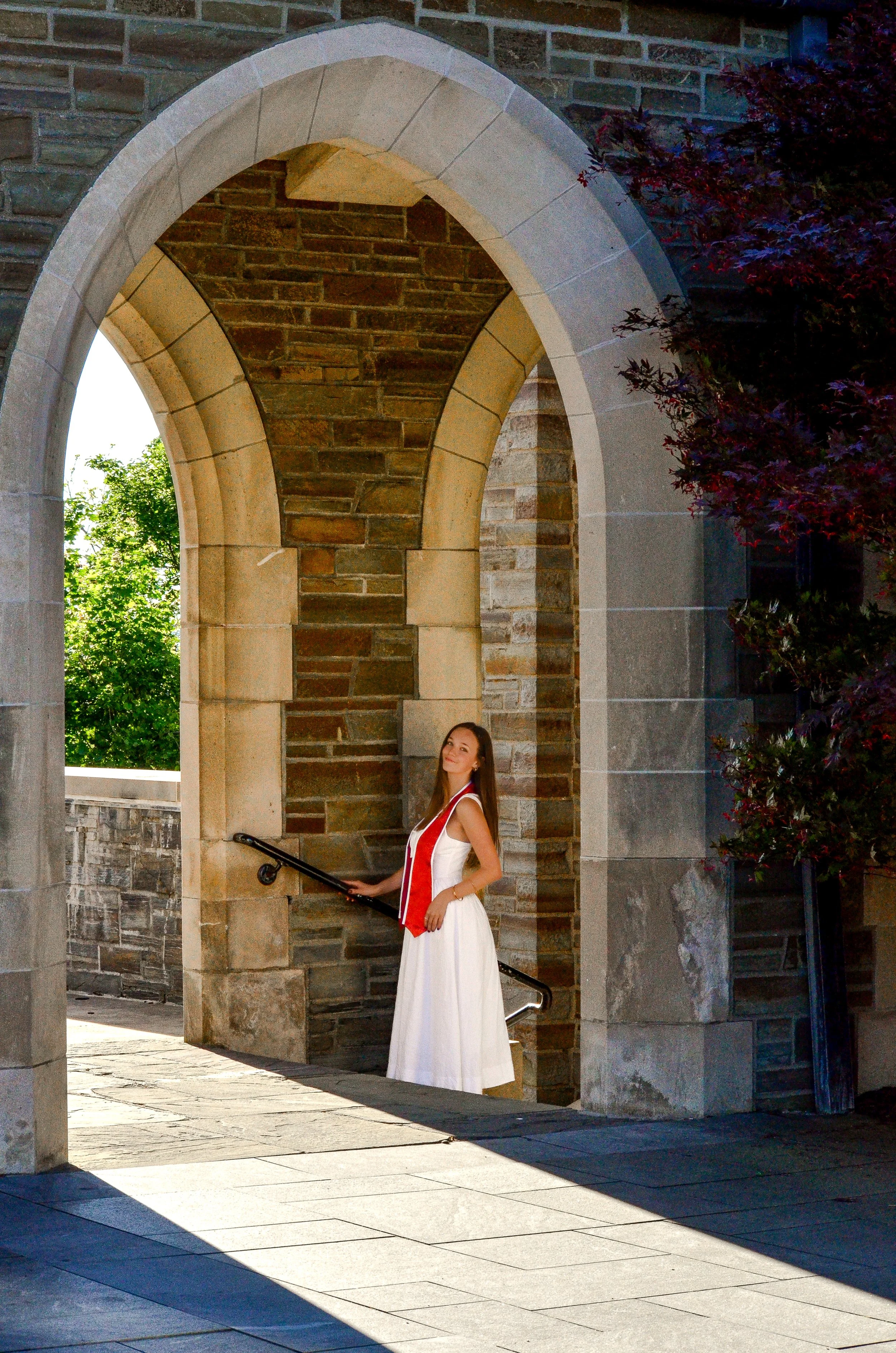 A woman in a white dress with a red stole stands under an arched stone structure, holding onto a black railing, with greenery in the background.