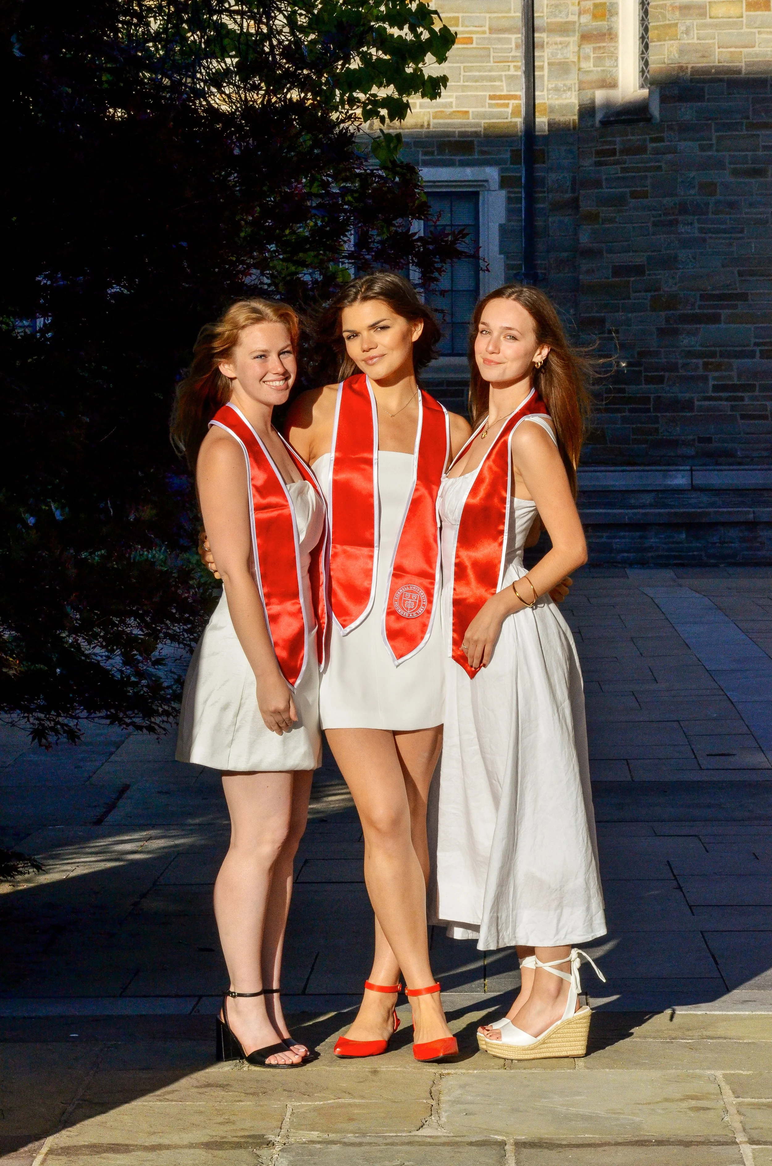 Three young women in graduation dresses and orange satin stoles standing outdoors on a stone path, posing for a photo.