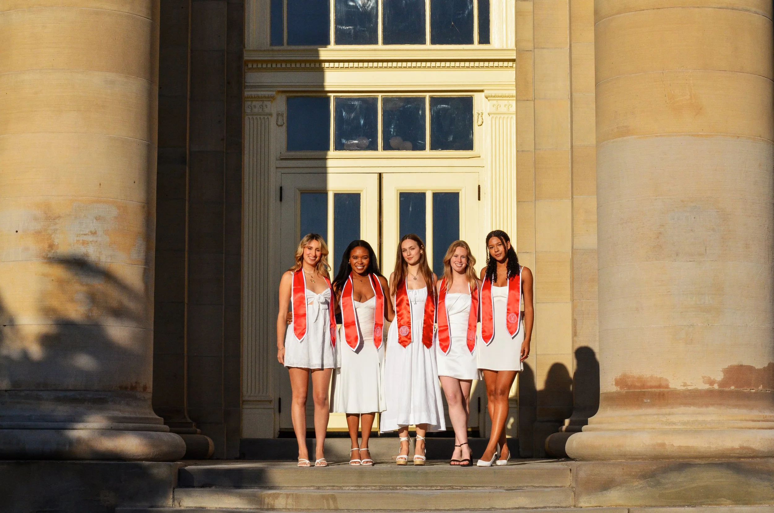 Five young women in white dresses wearing graduation stoles, standing on the steps in front of a building with large columns and a yellow door, smiling at the camera.