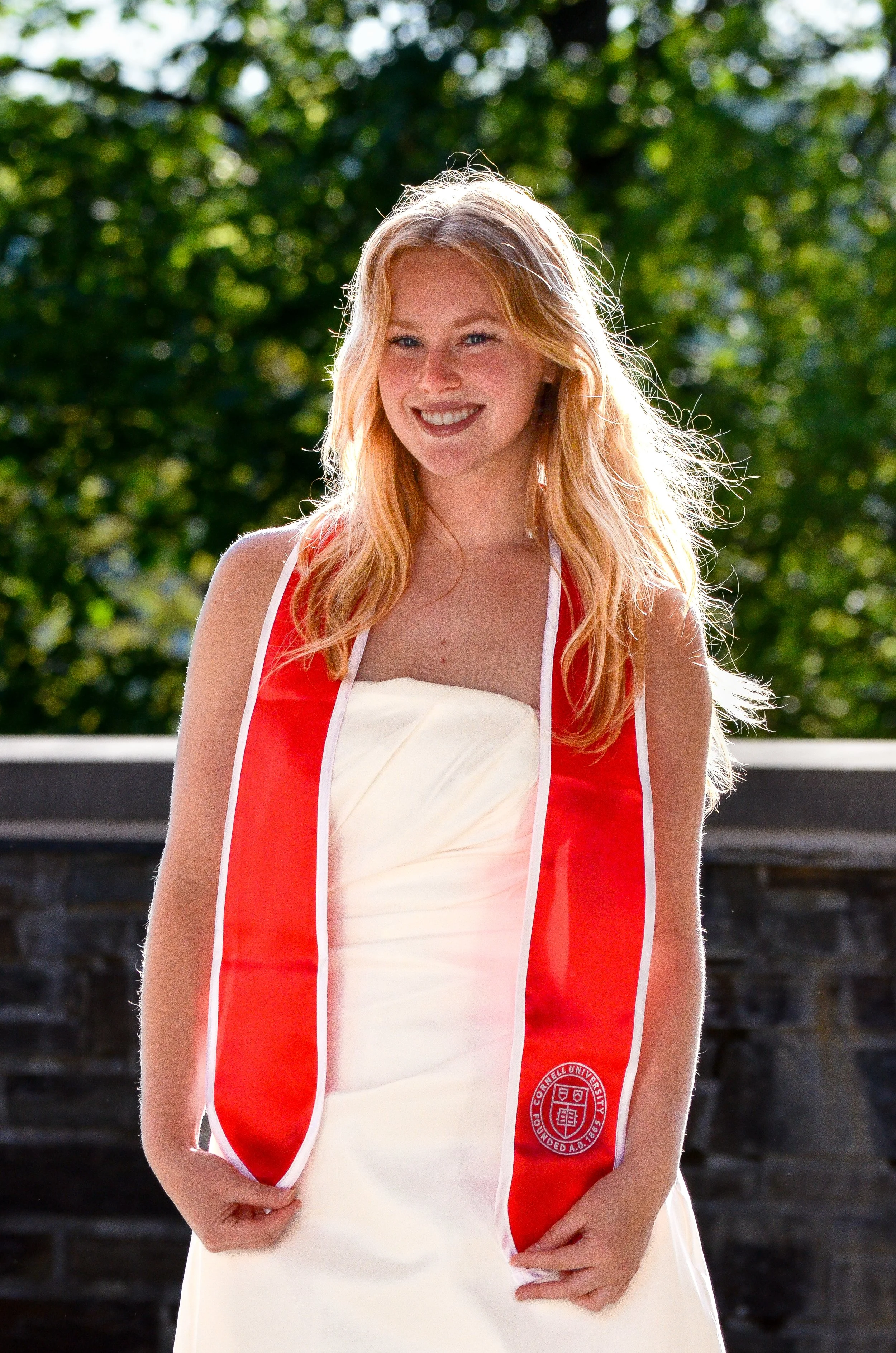 A young woman with long red hair wearing a white wedding dress and a red Cornell University honor cord. She is smiling and standing outdoors in front of a stone wall and green trees.