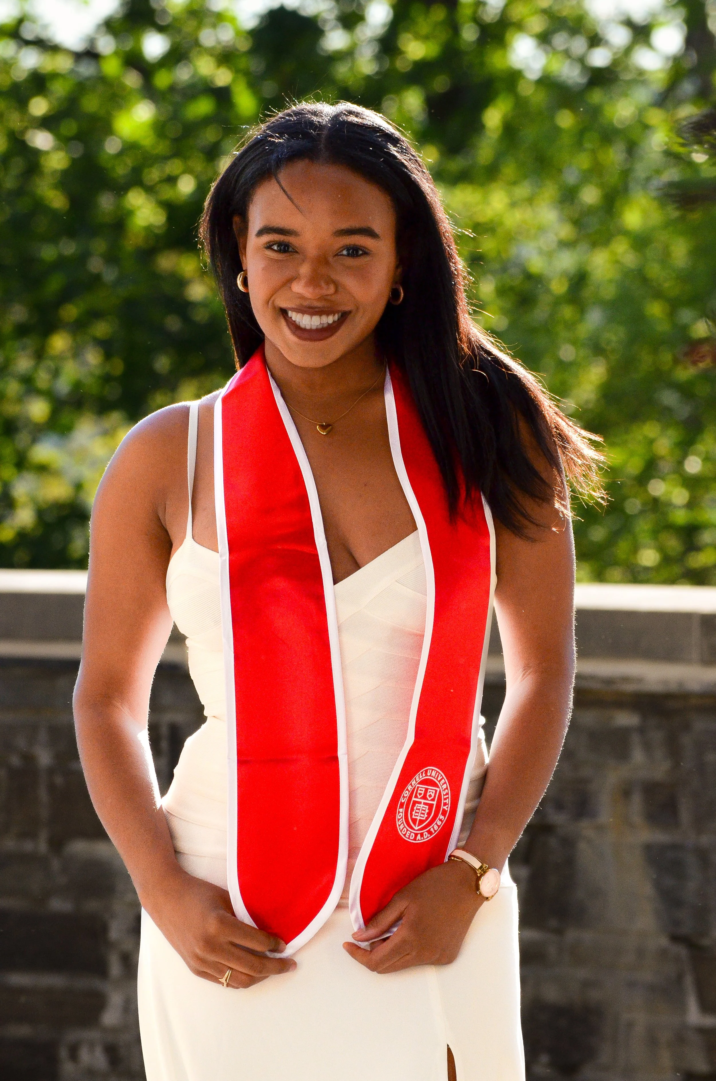 Young woman smiling outdoors, wearing a white dress and a red stole with a university emblem.
