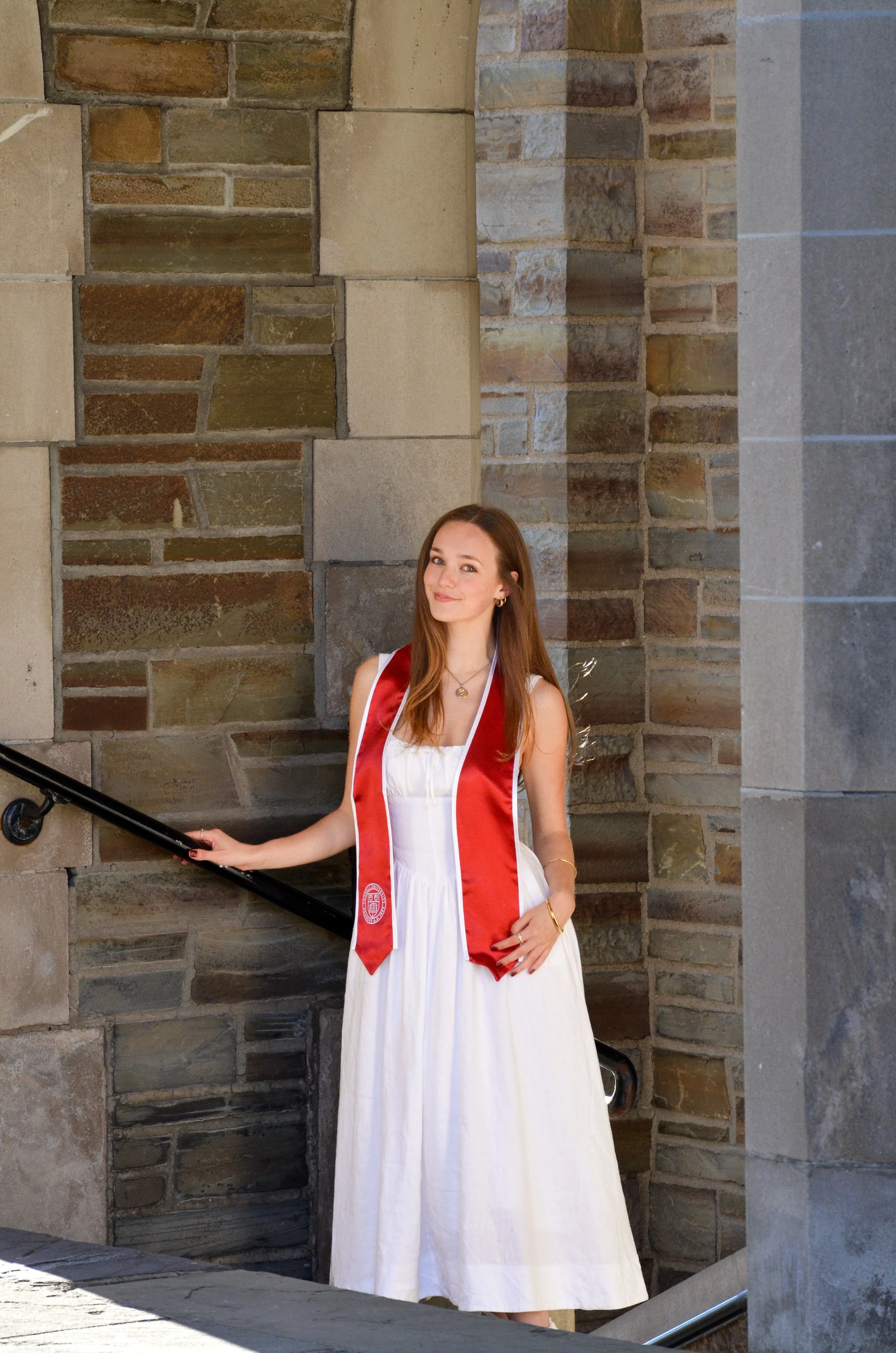 A young woman in a white dress with a red graduation stole, standing next to a stone wall staircase, smiling at the camera.