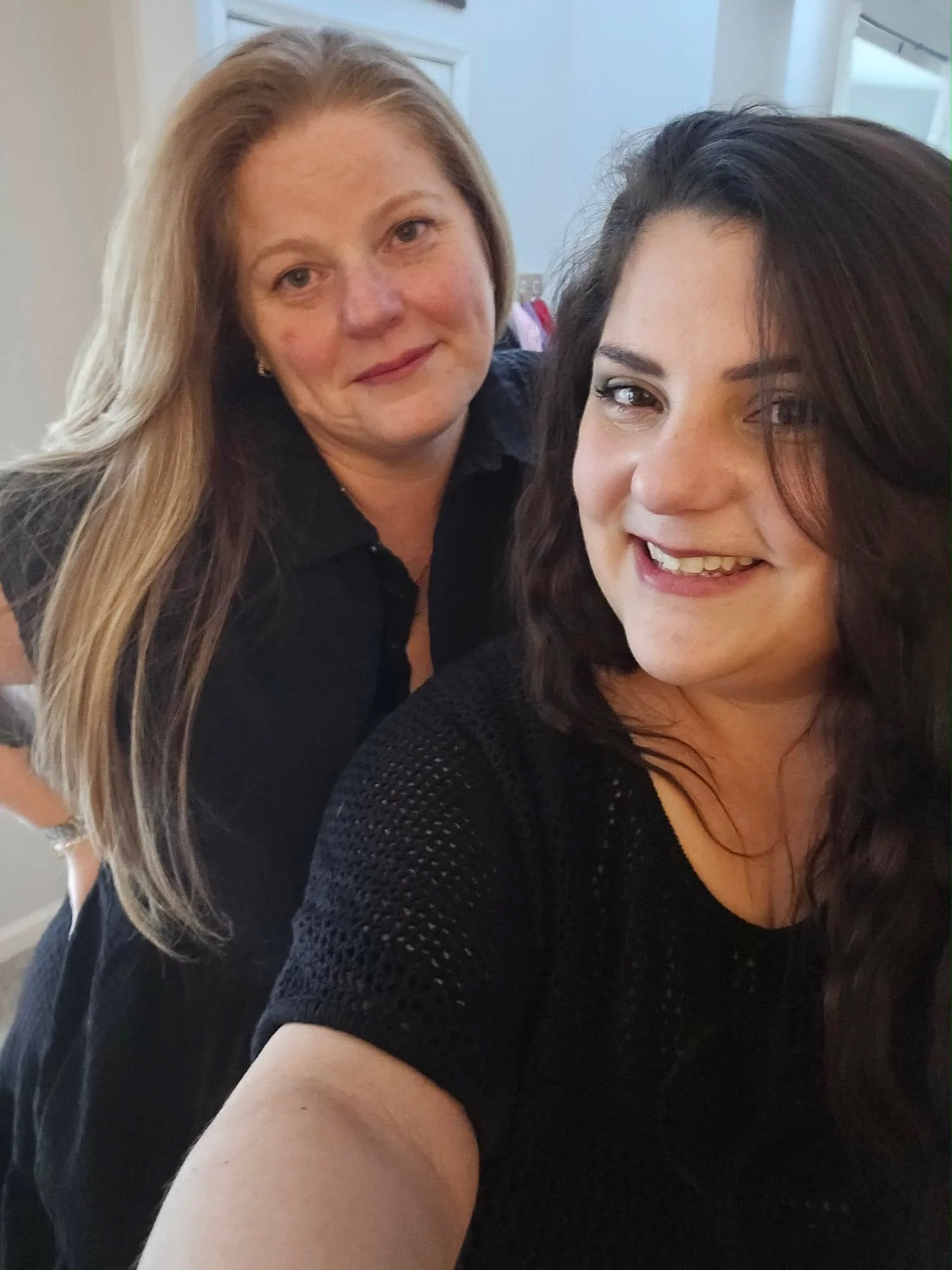 A woman with dark curly hair wearing a black knit top and a woman with long blonde hair wearing a black shirt standing close together, smiling at the camera.