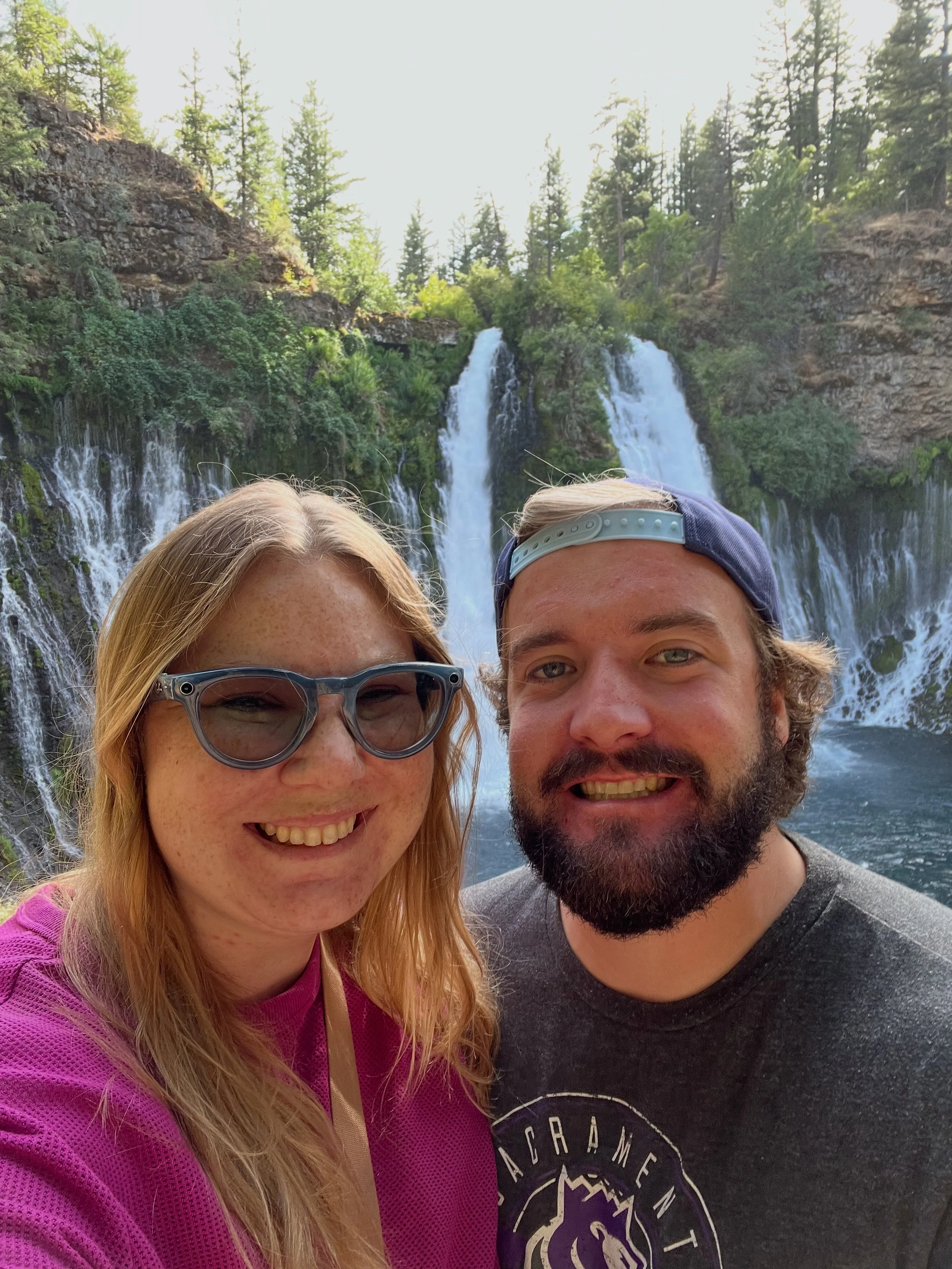 Smiling man and woman taking a selfie in front of a waterfall surrounded by trees.